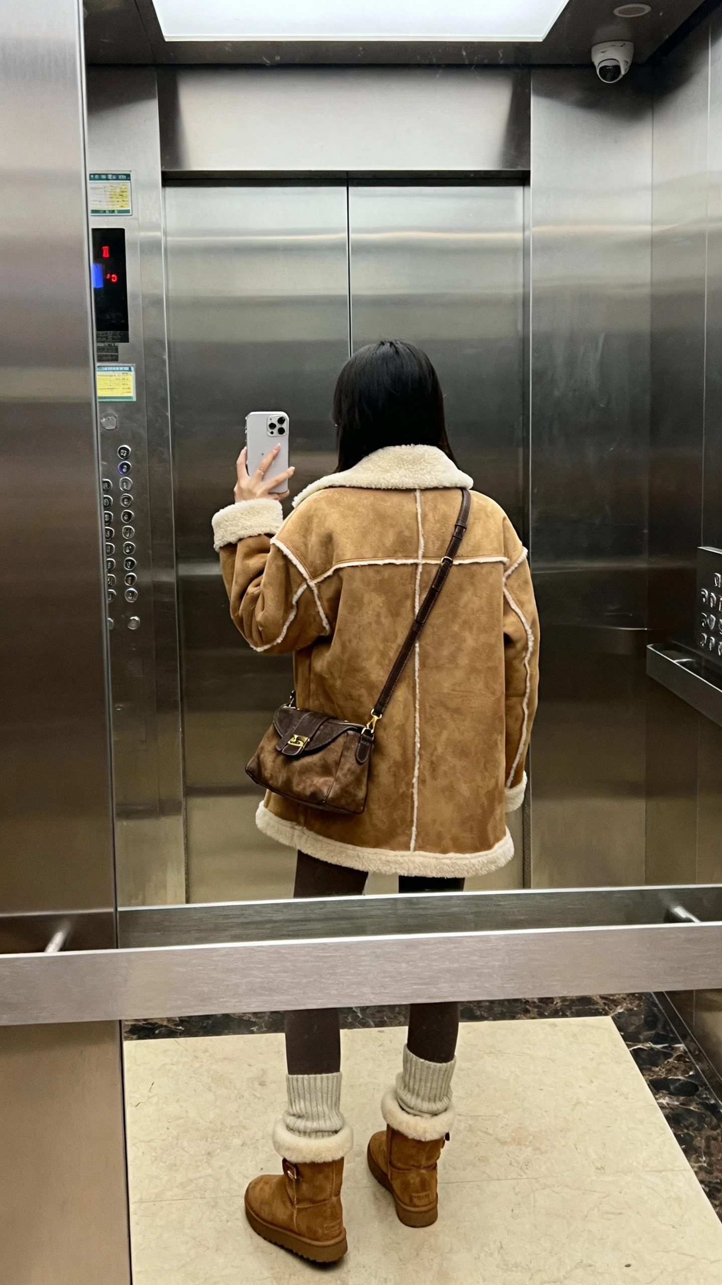 iPhone back-view selfie of a woman wearing a faux-shearling lined coat with matching shearling-lined boots and crossbody, cozy knit leggings, face not visible, elevator mirror, indoor lighting, natural iPhone photo quality.