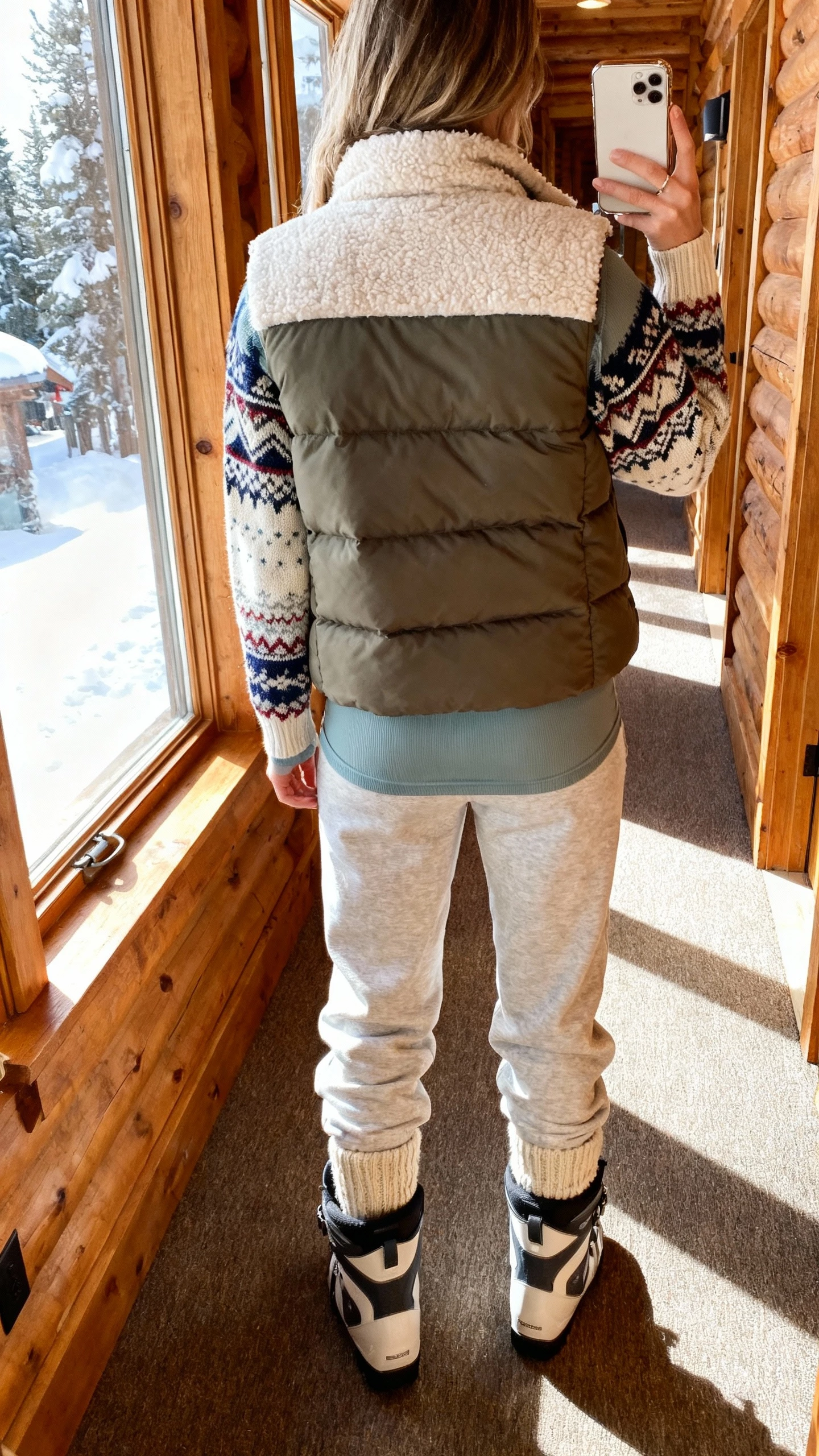 iPhone back-view selfie of a woman wearing a fleece-lined puffer vest over a thermal top, layered with a fair-isle sweater, soft joggers, wool socks, and après-ski boots; face not visible, ski-lodge style hallway with afternoon window light, casual iPhone photo.