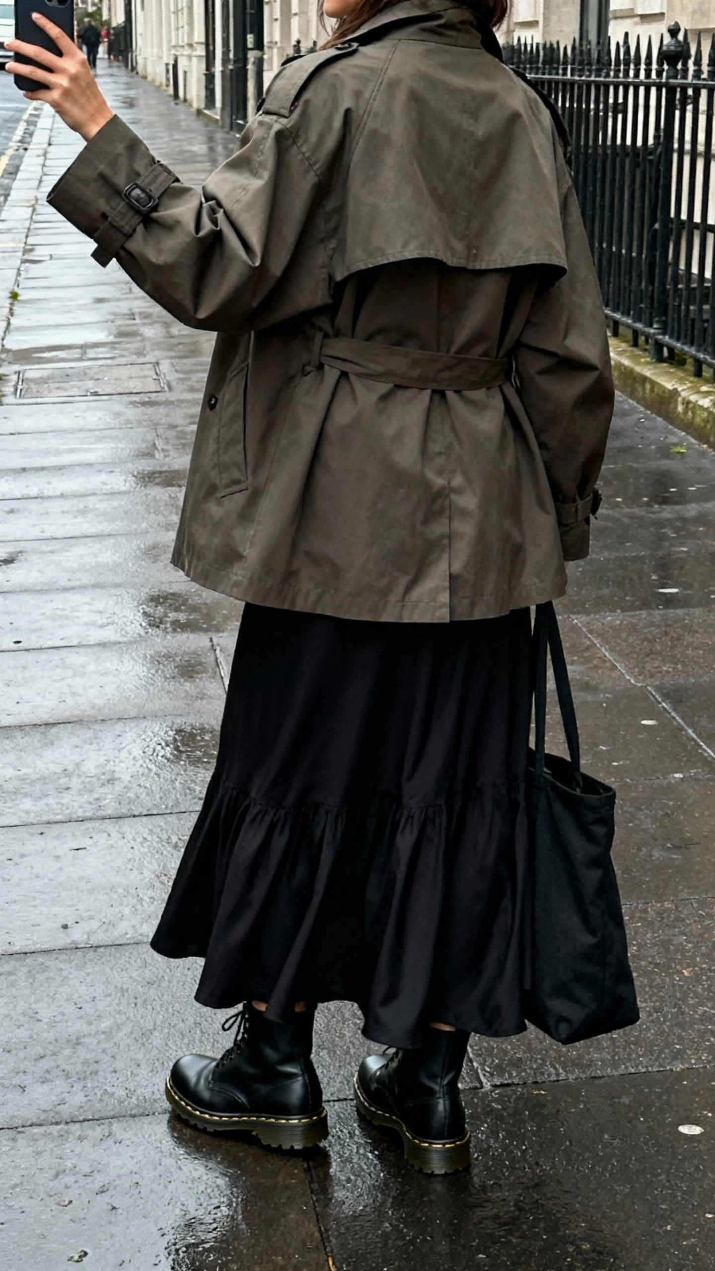 iPhone back-view selfie of a woman wearing a flowy black maxi skirt with an oversized trench coat, black Doc Martens, tote bag, face not visible, taken on a moody city sidewalk after rain, overcast daylight, casual iPhone aesthetic.