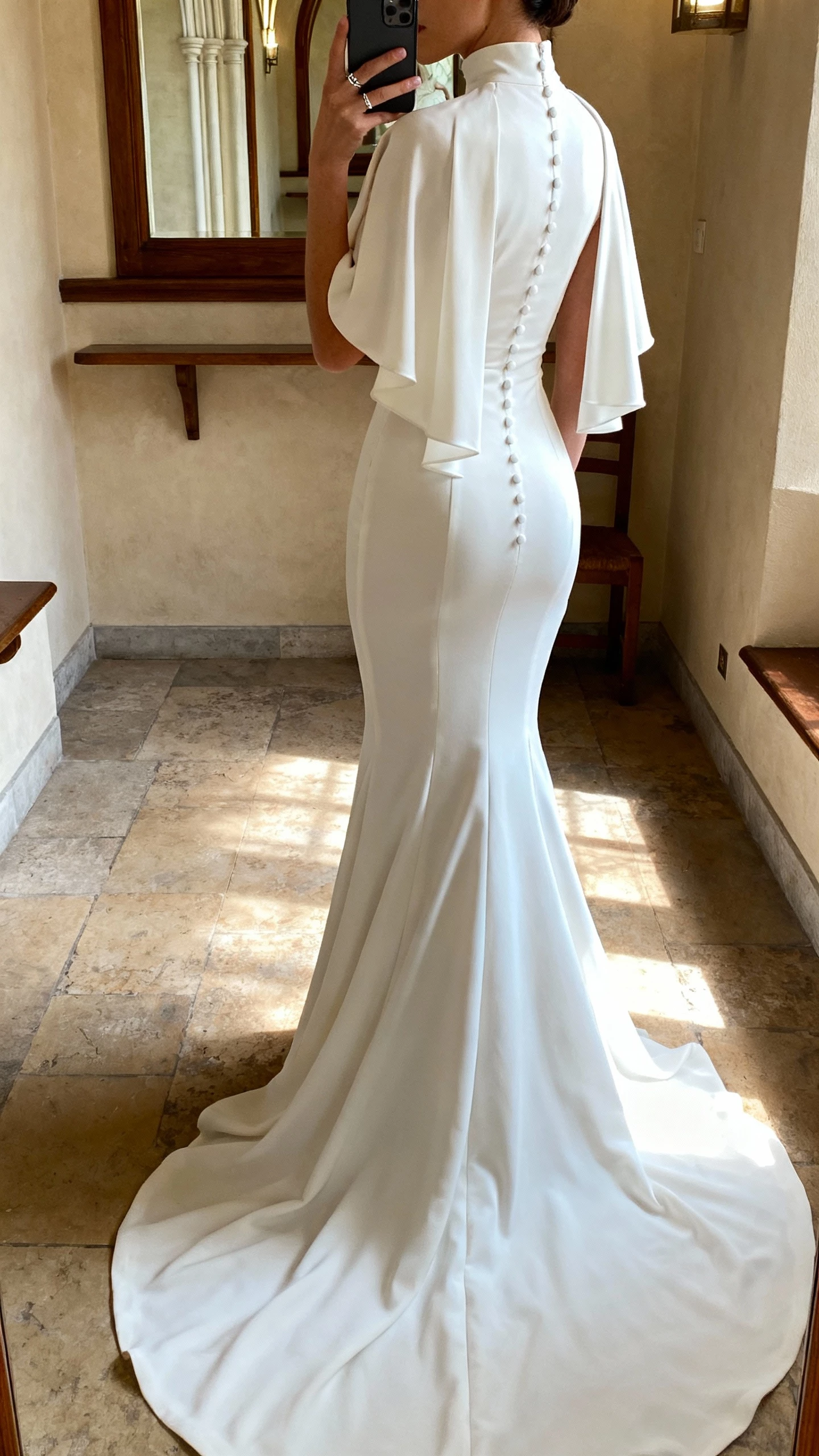 iPhone back-view selfie of a woman wearing a high-neck mermaid wedding dress with a dramatic cape back and buttons down the spine, minimal ring, face not visible, church vestibule mirror, gentle afternoon light, natural iPhone photo