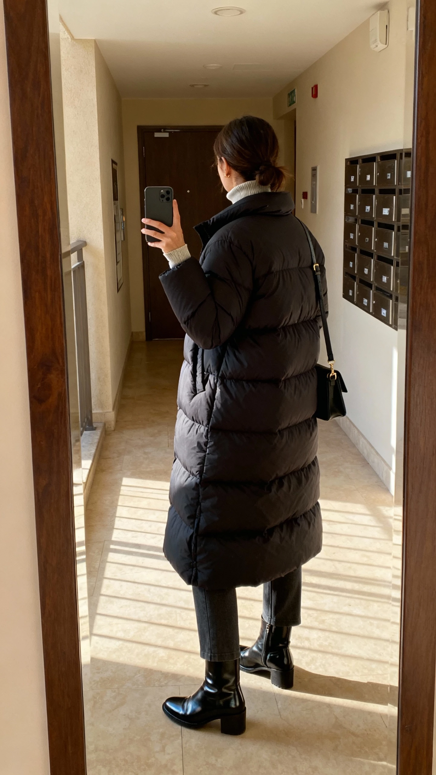 iPhone back-view selfie of a woman wearing a longline puffer over proportional layers (thin turtleneck and straight-leg pants), sleek winter boots, crossbody bag, face not visible, apartment lobby mirror, afternoon light, natural iPhone quality.