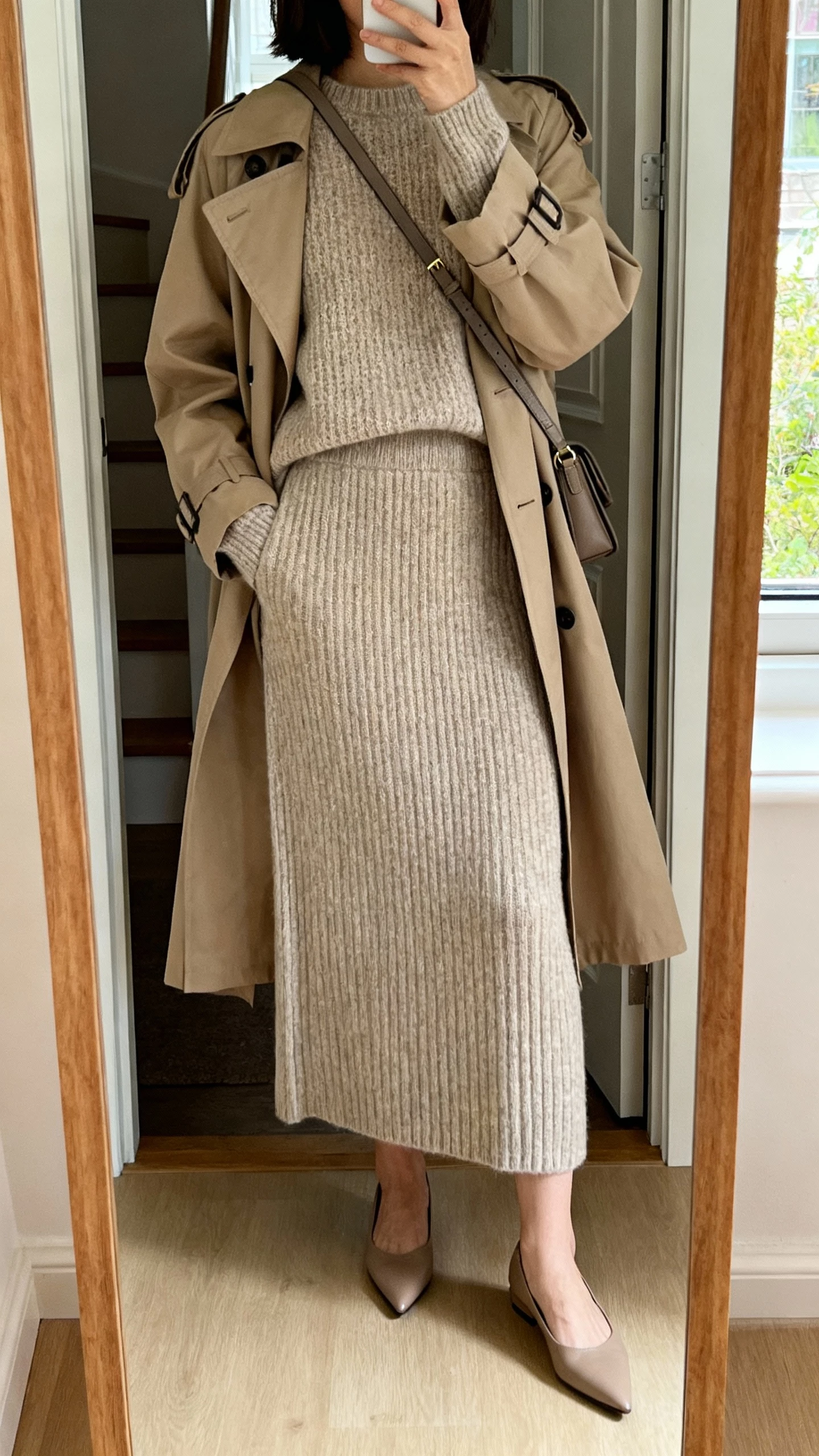 iPhone back-view selfie of a woman wearing a matching knit set (fine-knit sweater and ribbed knit skirt) topped with a classic trench coat, pointed flats and a crossbody, face not visible, apartment entryway mirror, daylight, natural iPhone photo.