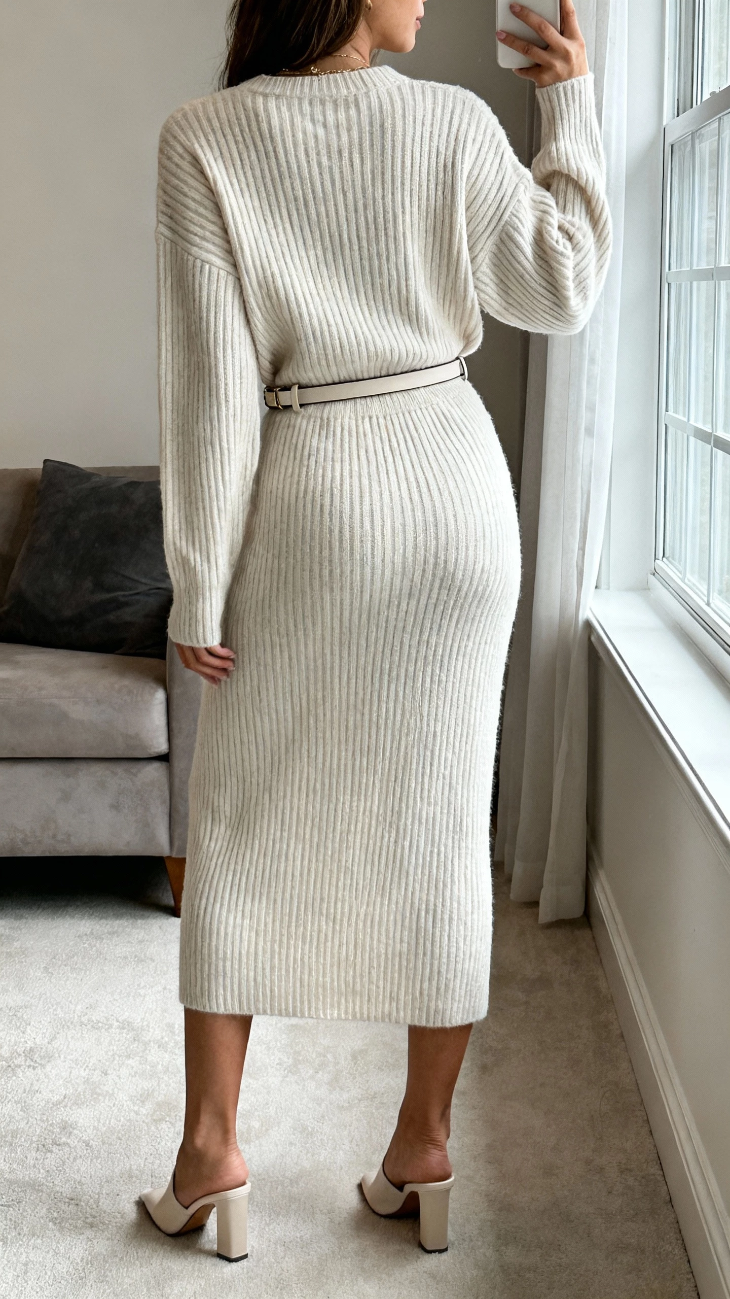 iPhone back-view selfie of a woman wearing a monochrome knit set (ribbed sweater and matching knit skirt) with heeled mules and a slim belt, face not visible, living room by a window, natural iPhone photo quality