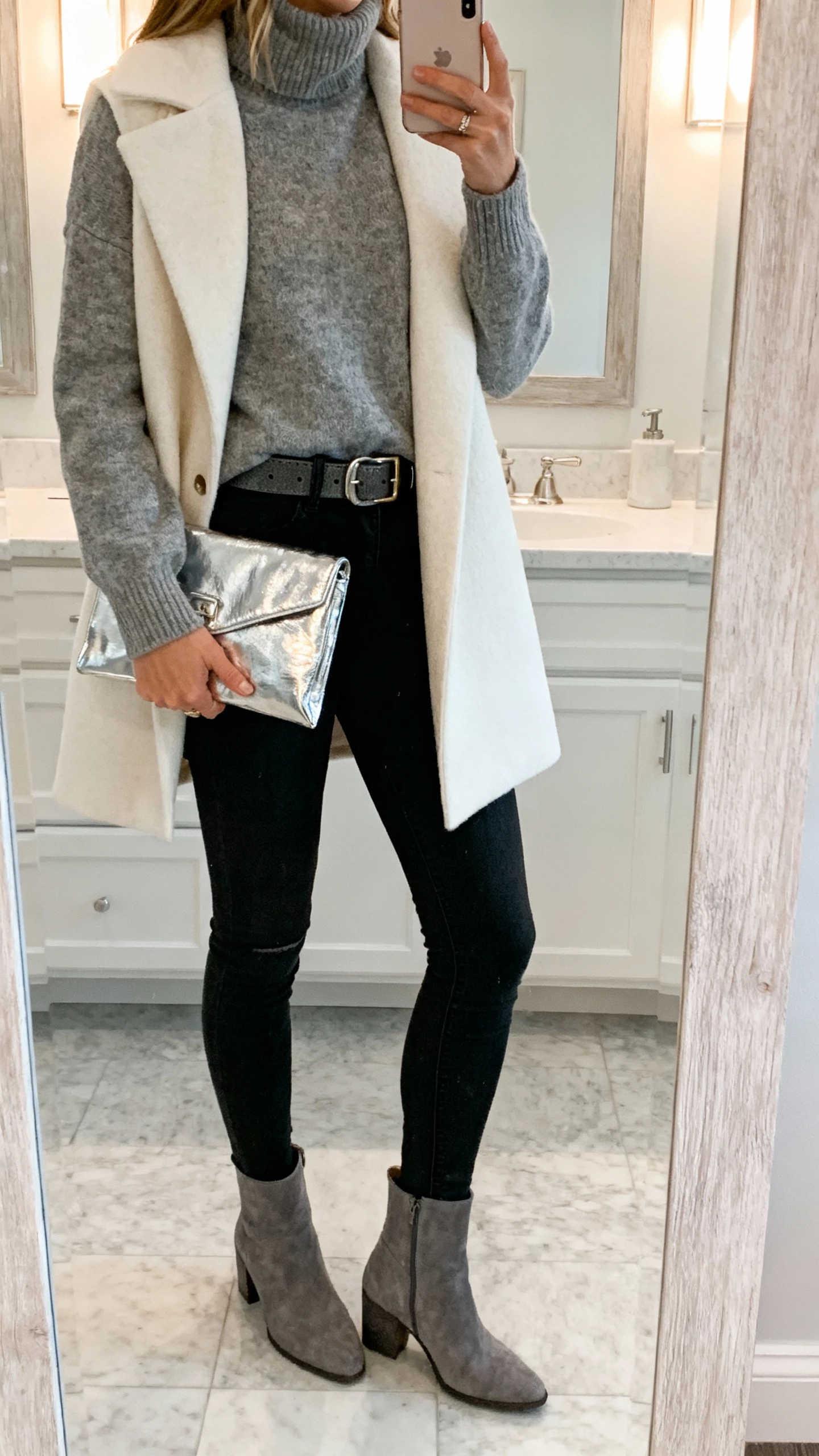 iPhone back-view selfie of a woman wearing a pewter sweater, ivory coat, black skinny jeans, metallic molten silver clutch and belt, gray ankle boots, face not visible, bathroom mirror with soft overhead light, casual iPhone photo quality