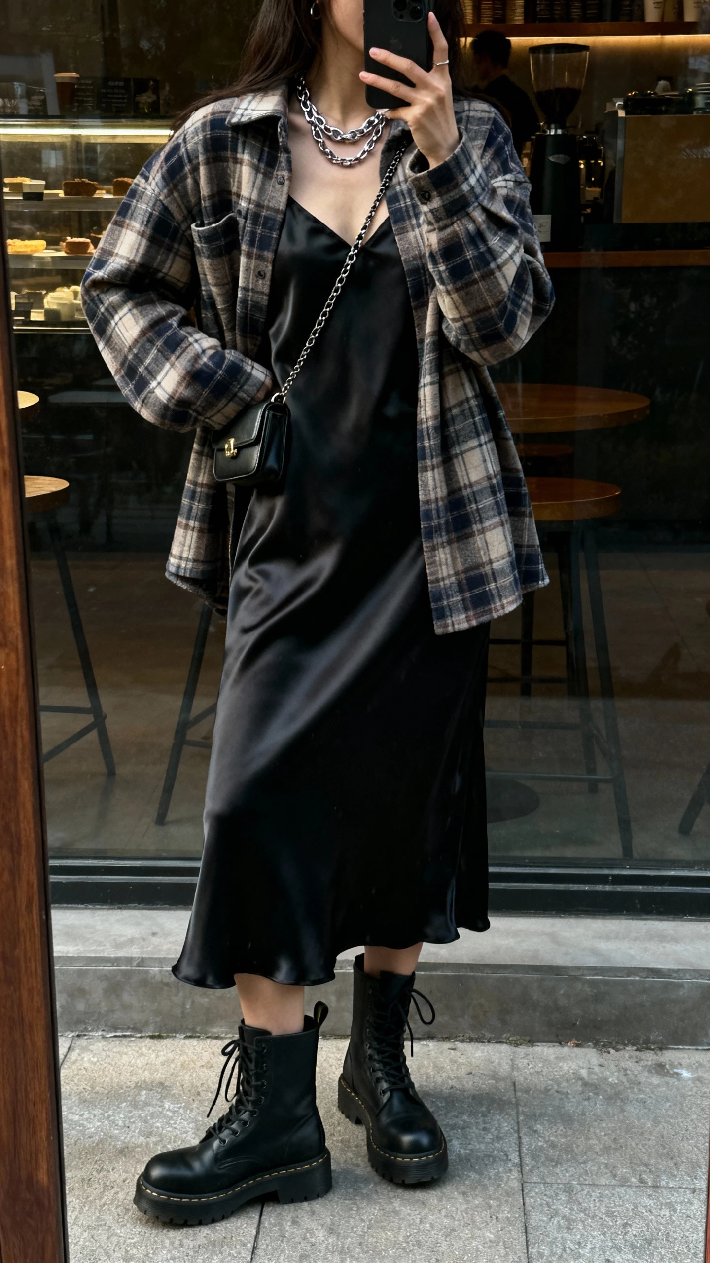 iPhone back-view selfie of a woman wearing a plaid shacket over a silky black slip dress, combat boots, layered chain necklace, and a small crossbody, face not visible, taken in a dim coffee shop window reflection with afternoon natural light, natural iPhone photo quality.