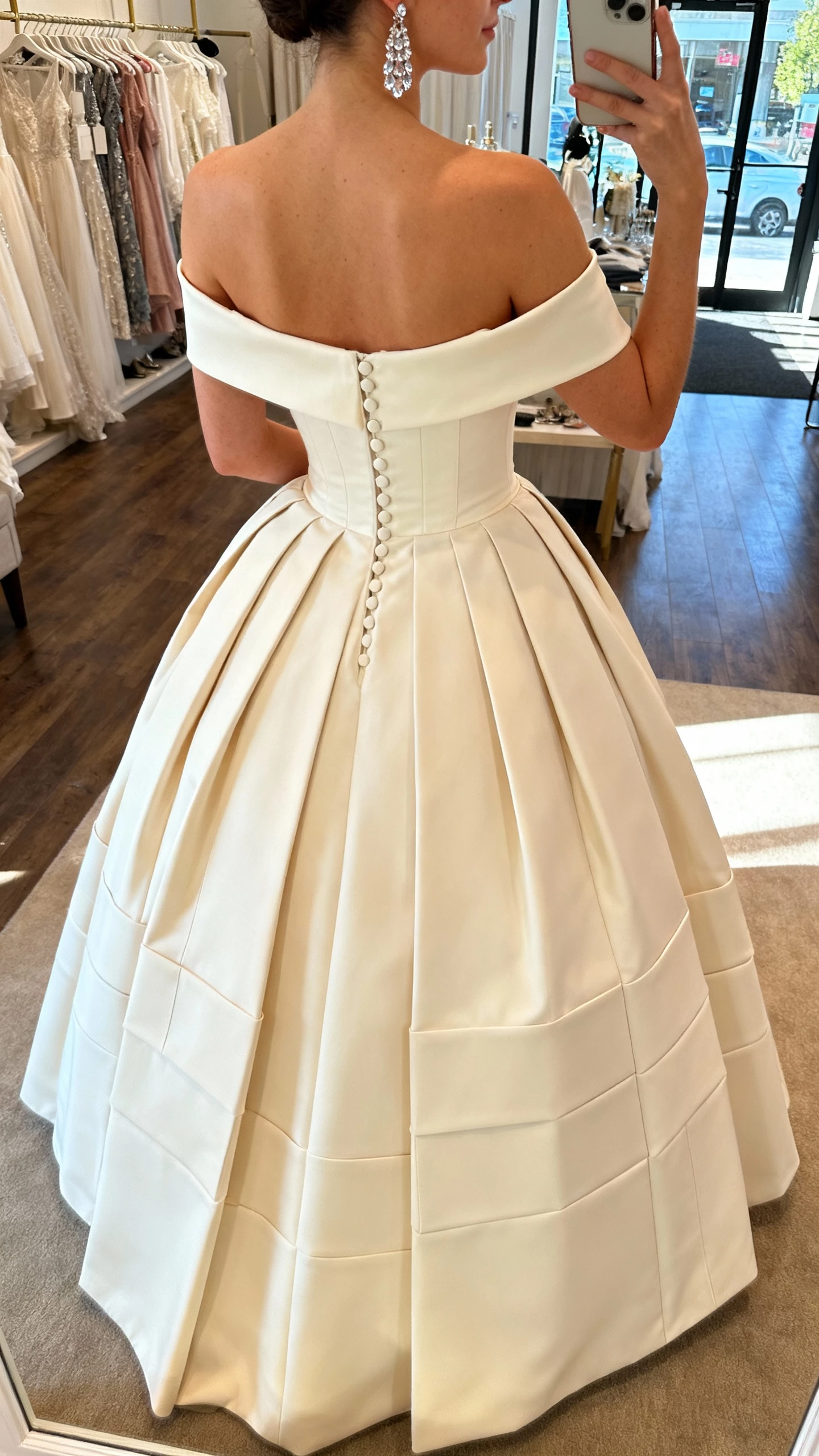 iPhone back-view selfie of a woman wearing a regal mikado ballgown with structured box pleats, off-the-shoulder neckline, covered buttons down the back, crystal drop earrings, face not visible, boutique fitting room, overhead warm light, natural iPhone photo quality