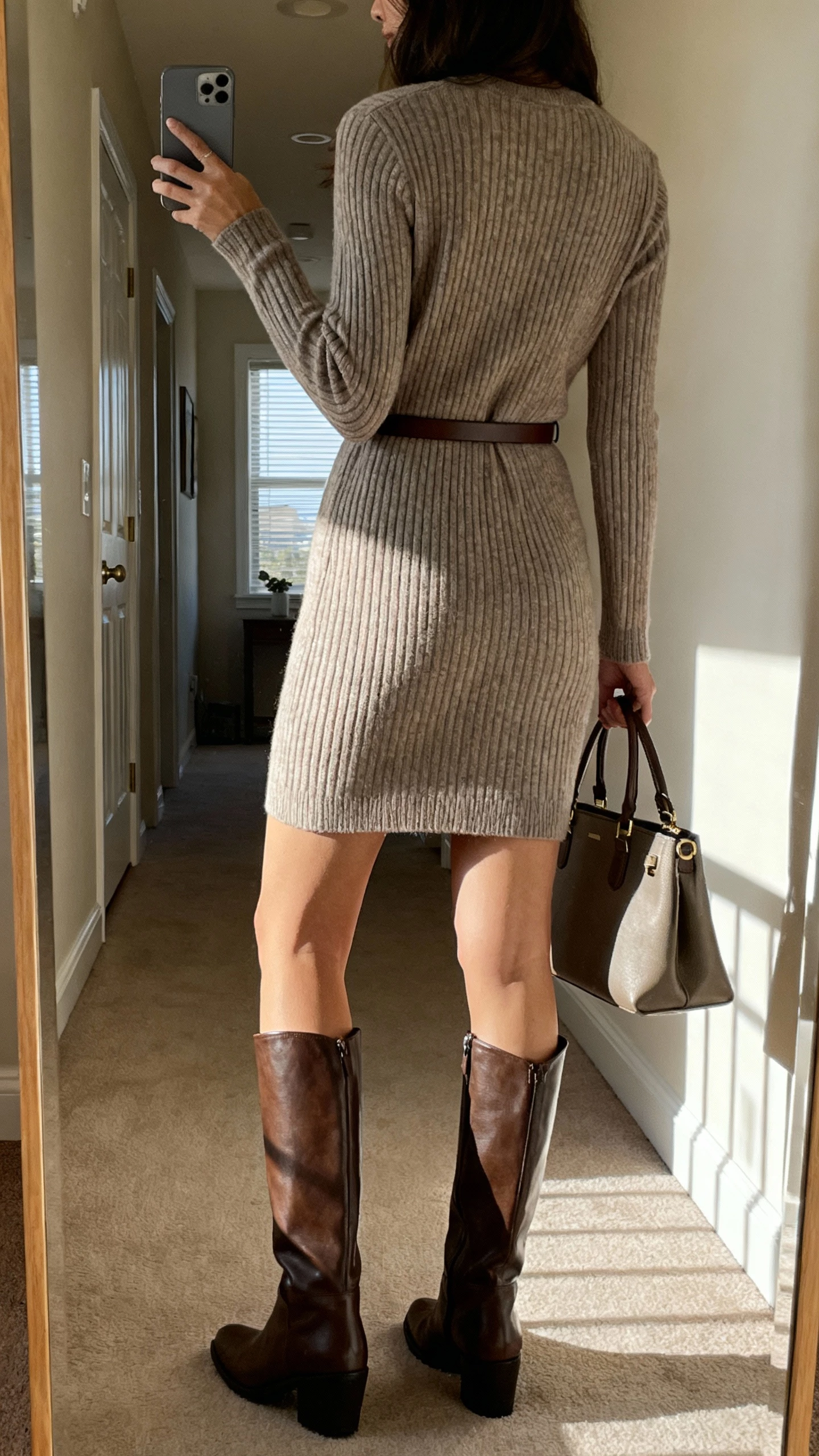 iPhone back-view selfie of a woman wearing a ribbed knit dress with knee boots and a slim belt, holding a structured tote, face not visible, apartment hallway mirror, afternoon window light, natural iPhone quality.