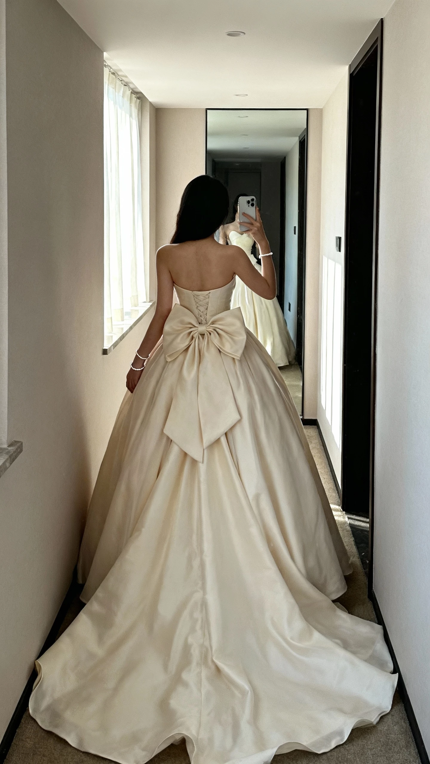 iPhone back-view selfie of a woman wearing a romantic ball gown with an oversized bow at the back, flowing train, sweetheart bodice, and dainty bracelet, face not visible, corridor mirror, morning window light, casual iPhone quality
