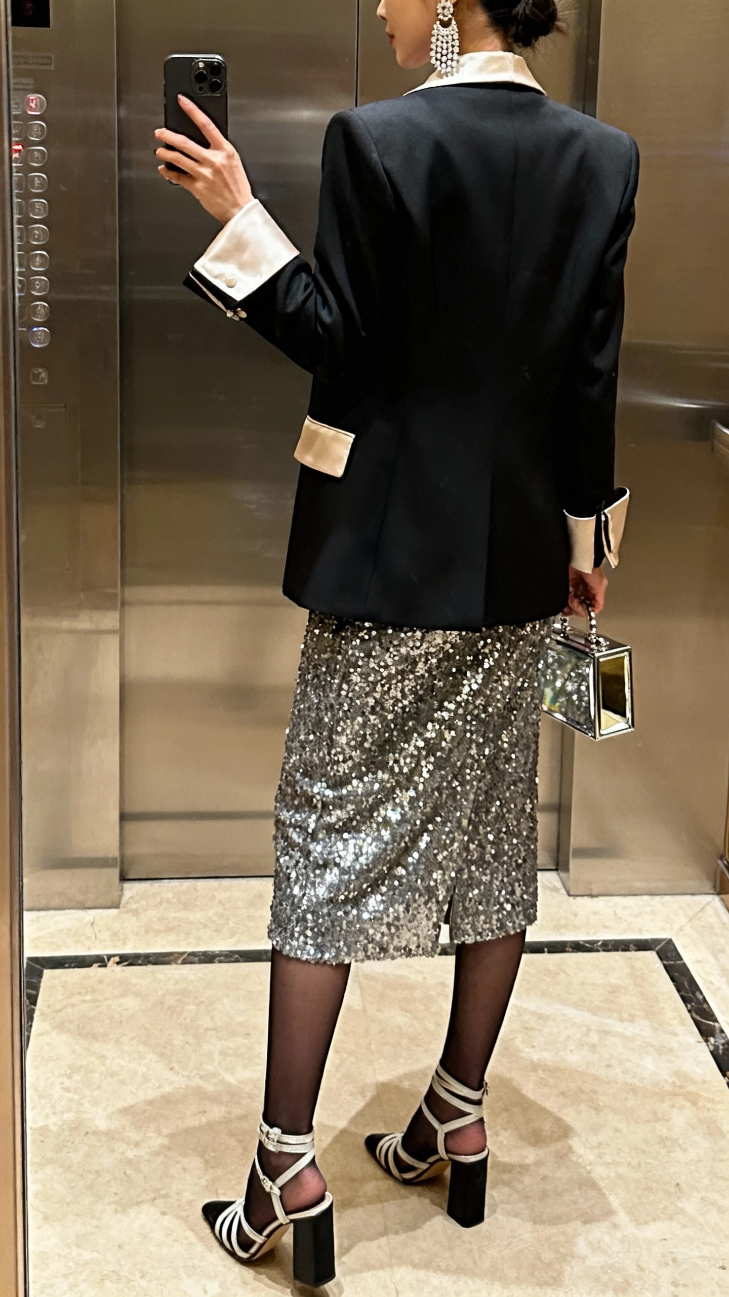 iPhone back-view selfie of a woman wearing a sequin midi dress with a sharp tuxedo blazer (satin lapels), strappy block heels, chandelier earrings, a small mirrored box clutch, and sheer black tights; face not visible, elevator mirror, warm indoor lighting, natural iPhone quality.