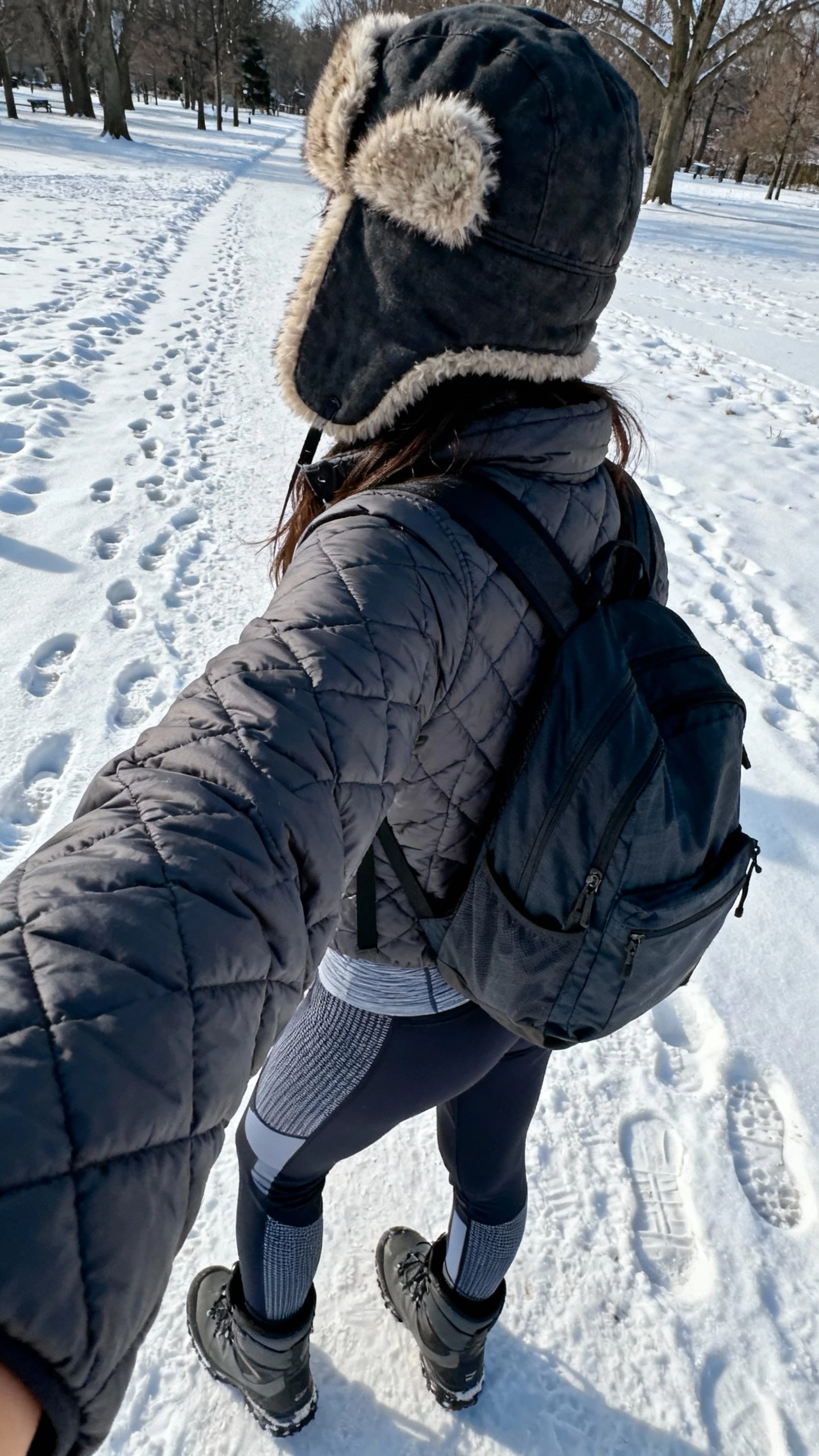 iPhone back-view selfie of a woman wearing a shearling-lined trapper hat, quilted parka, thermal base layer, technical hiking pants, waterproof boots, and a nylon backpack; face not visible, snowy park trail, cool daylight, casual iPhone photo.