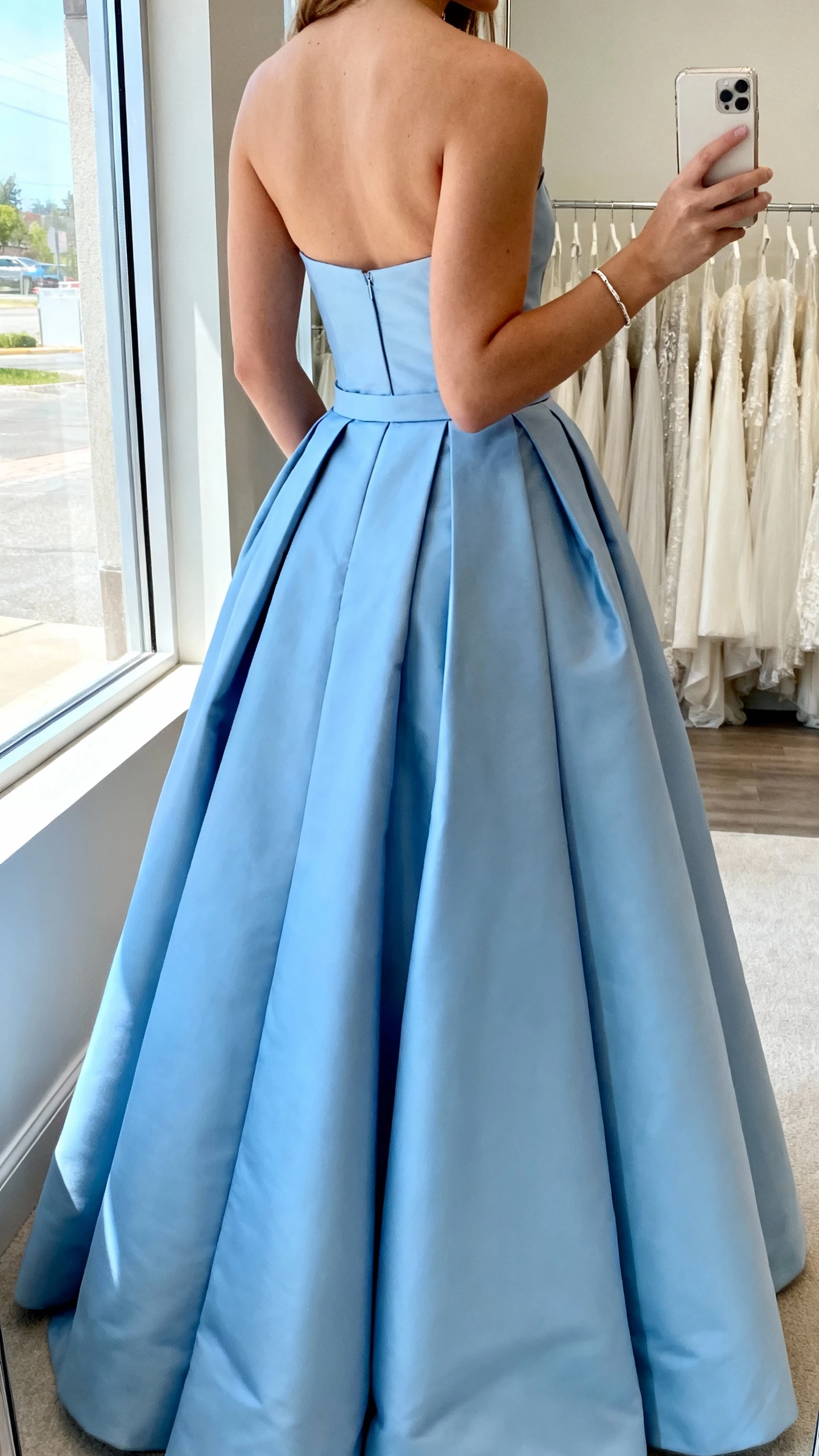 iPhone back-view selfie of a woman wearing a soft blue ballgown with structured skirt, simple bracelet, face not visible, bridal boutique mirror, daylight from window, casual iPhone shot
