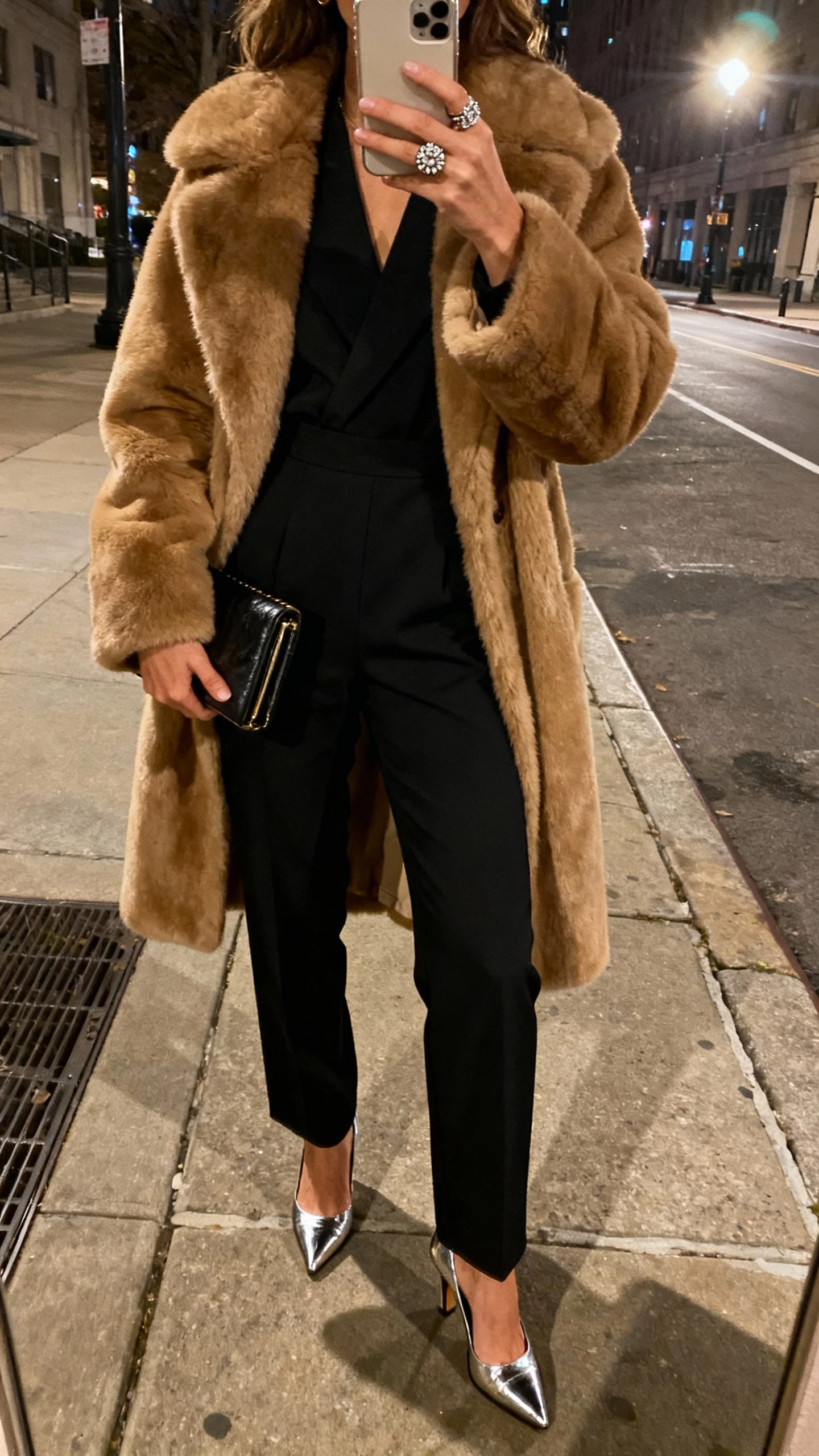 iPhone back-view selfie of a woman wearing a tailored black jumpsuit under a tan faux-fur coat, metallic heels, clutch, statement ring, face not visible, city sidewalk at night, streetlight glow, casual iPhone aesthetic