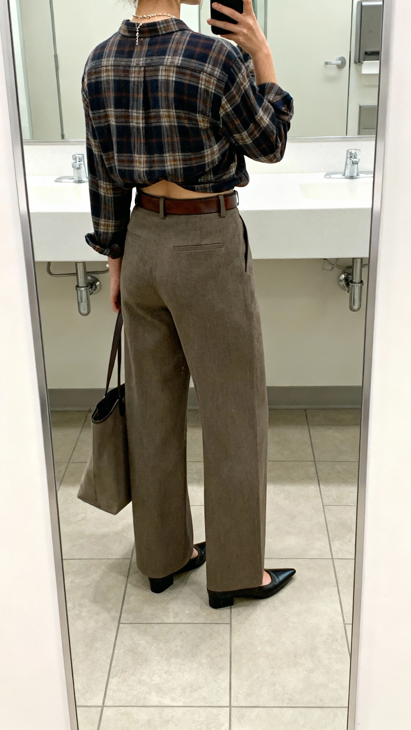 iPhone back-view selfie of a woman wearing a tailored plaid flannel shirt half-tucked into high-waisted wide-leg trousers, leather belt, pointed loafers, delicate necklace, tote bag, face not visible, office restroom mirror with bright indoor light, natural iPhone aesthetic.