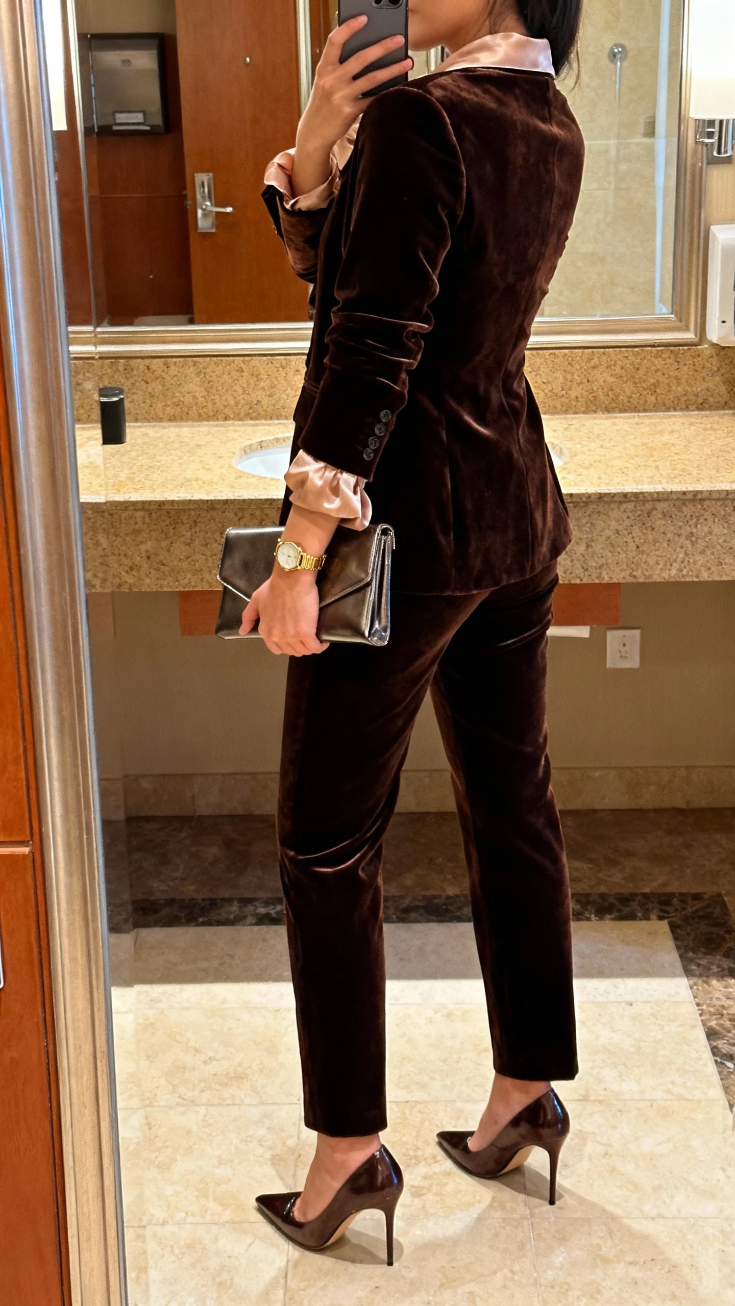 iPhone back-view selfie of a woman wearing a velvet power suit with a soft silk blouse, heeled pumps, sleek clutch, subtle gold watch, face not visible, hotel restroom mirror, warm indoor lighting, casual iPhone shot.