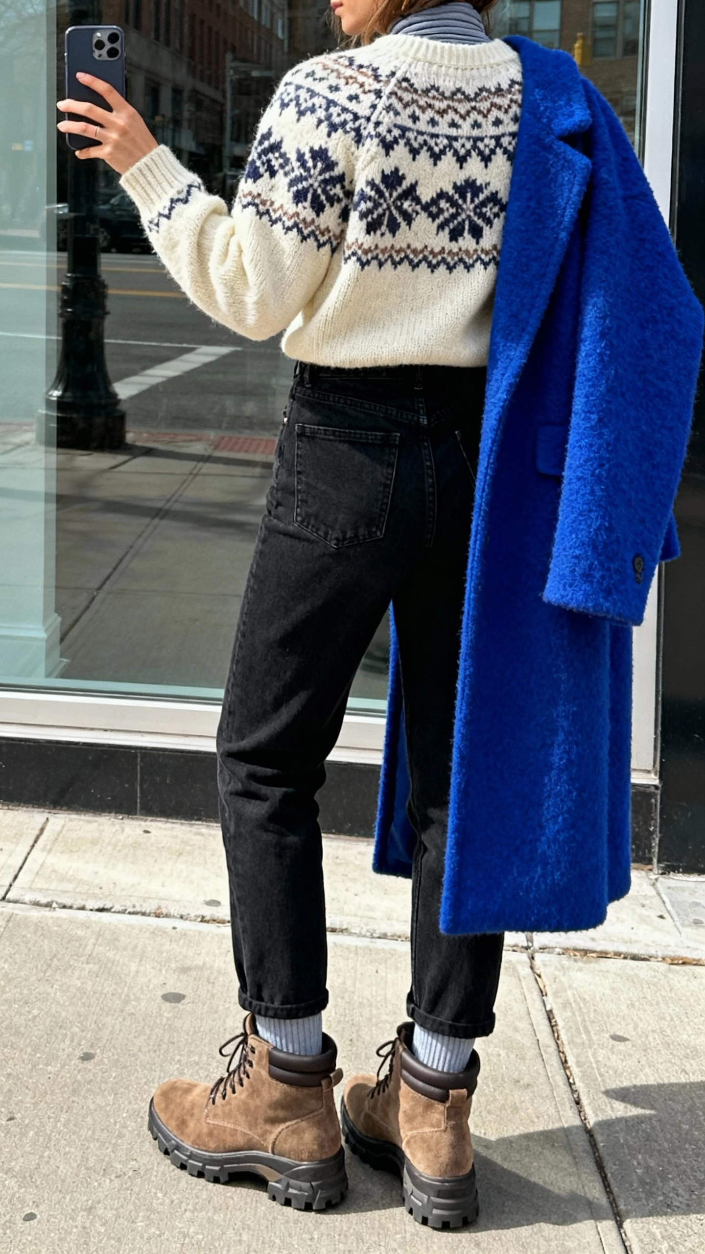 iPhone back-view selfie of a woman wearing an ivory Fair Isle sweater, high-waisted black coated jeans, chunky suede hiker boots, bold cobalt wool coat, thermal base layer underneath, face not visible, taken on a city sidewalk window reflection in daylight, casual iPhone photo.