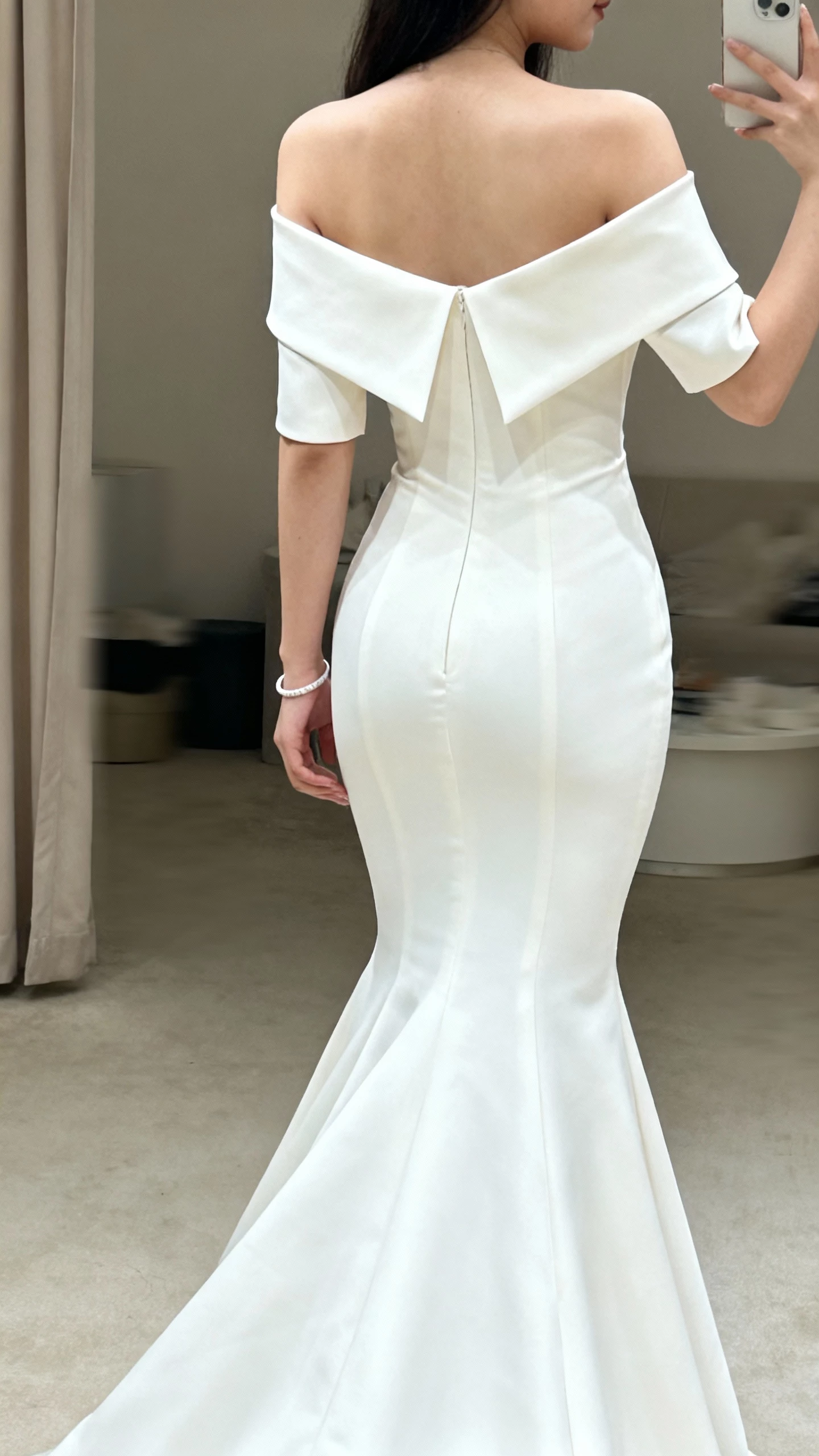 iPhone back-view selfie of a woman wearing an off-the-shoulder mermaid wedding gown with a sharp architectural neckline and structured sleeves, fitted skirt flaring at knees, simple bracelet, face not visible, bridal boutique fitting room lighting, casual iPhone aesthetic