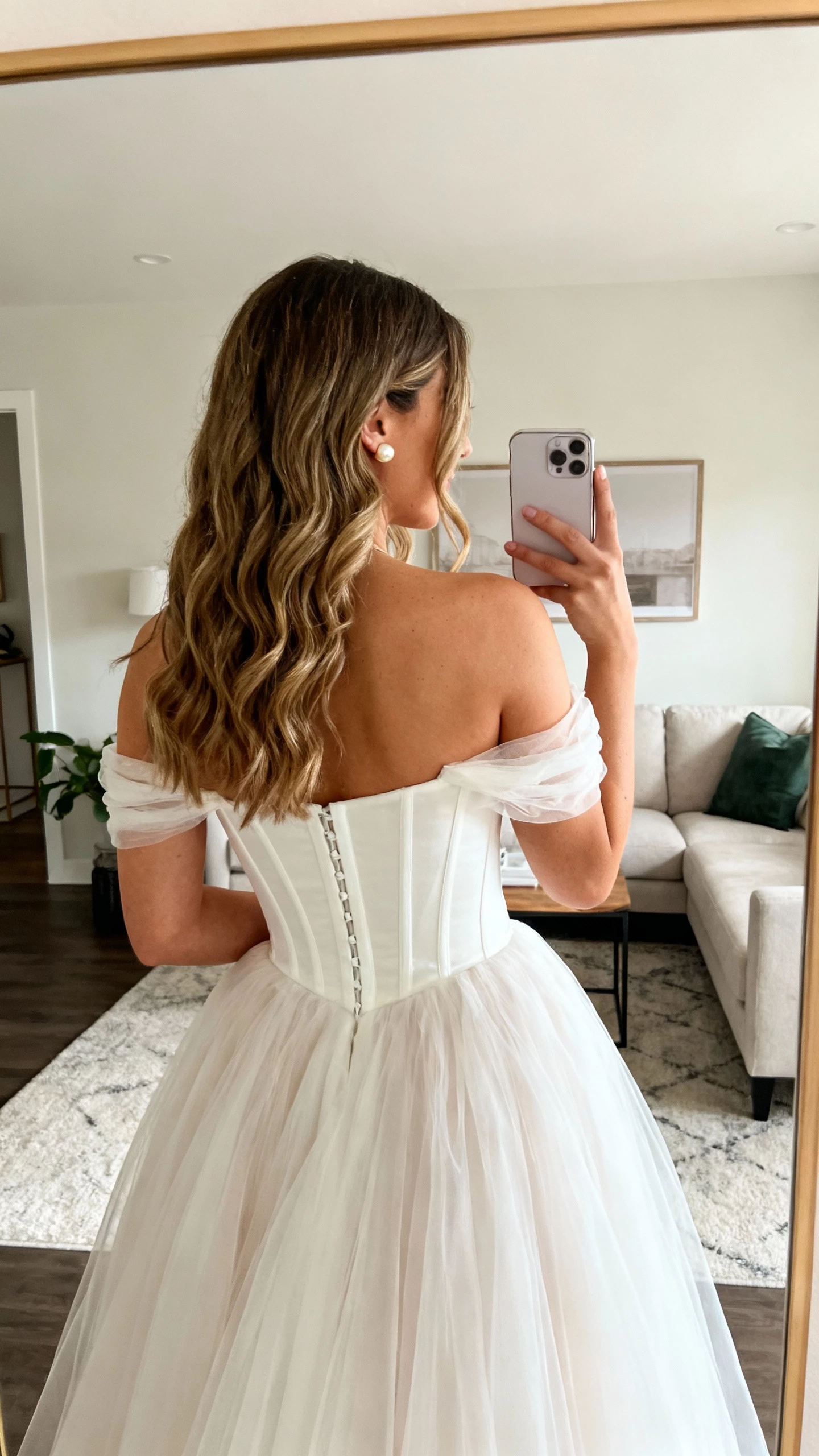 iPhone back-view selfie of a woman wearing an off-the-shoulder wedding dress that frames the collarbone, fitted corset bodice, romantic tulle skirt, pearl studs, loose waves over shoulders, face not visible, living room mirror, soft daylight, casual iPhone photo.