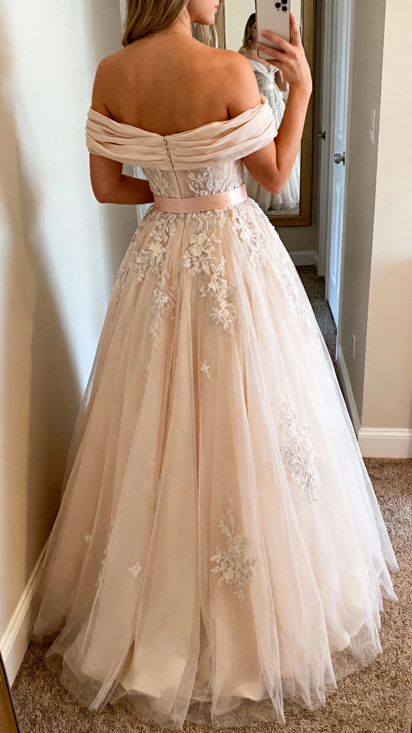 iPhone back-view selfie of a woman wearing an off-shoulder fairytale ball gown with soft draped neckline, romantic tulle layers, delicate lace appliqués, and a subtle waist sash, face not visible, hallway mirror, warm indoor lighting, casual iPhone shot