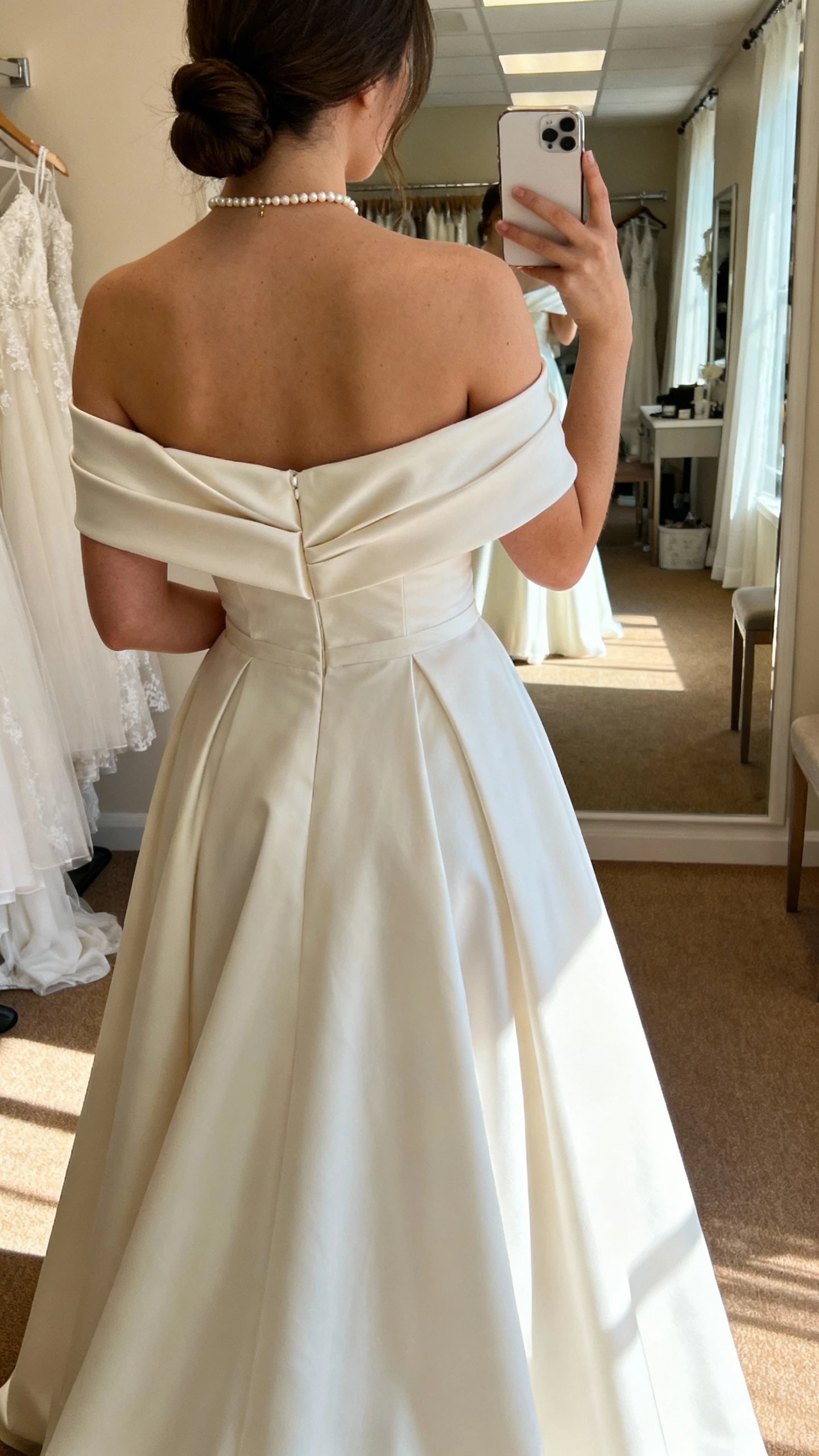 iPhone back-view selfie of a woman wearing an off-shoulder A-line wedding gown with folded neckline, flowing skirt, dainty pearl necklace, and low bun; face not visible, bridal suite mirror, warm afternoon light, casual iPhone photo.
