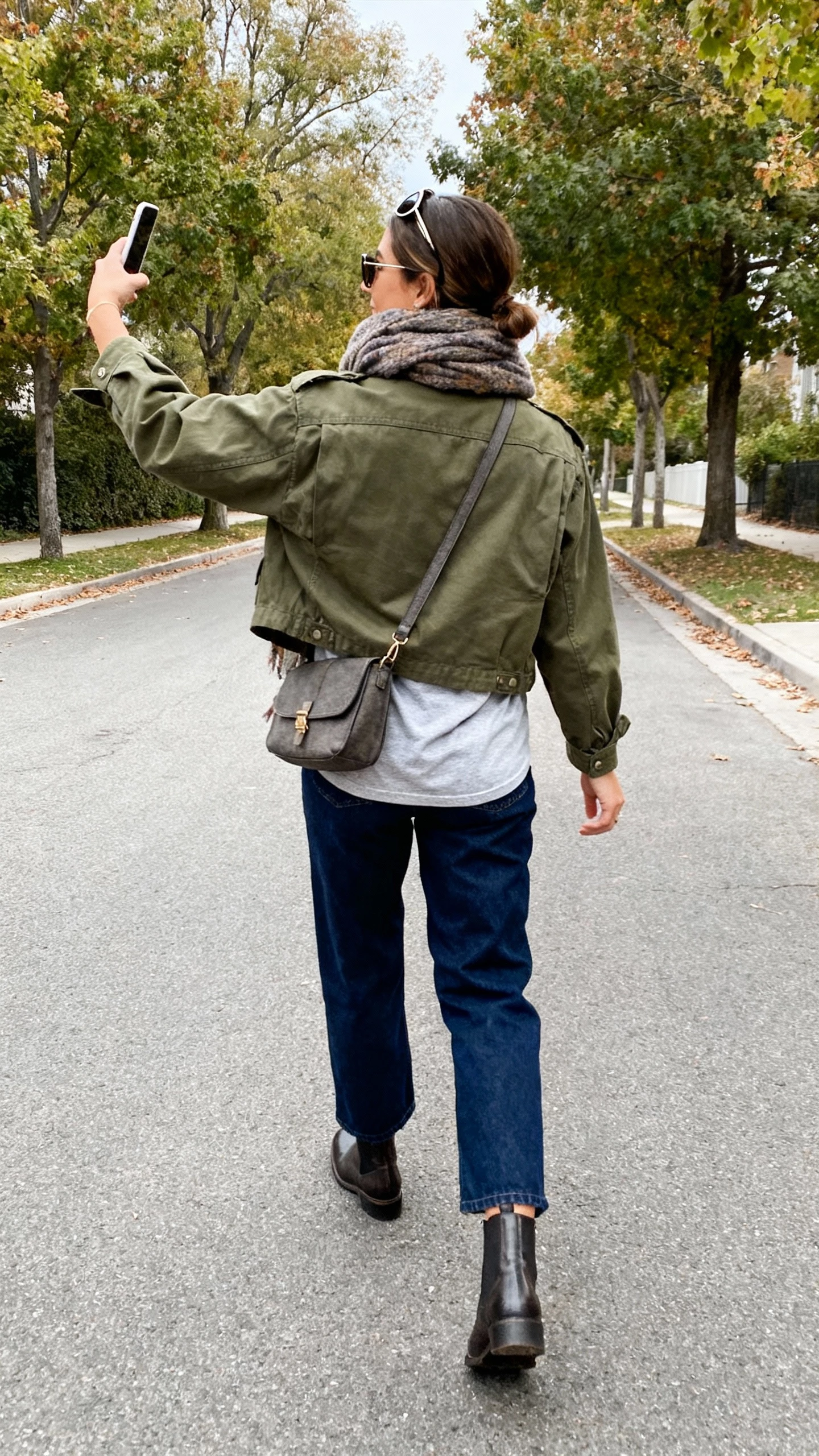 iPhone back-view selfie of a woman wearing an olive utility jacket over a soft tee, dark blue relaxed jeans, Chelsea boots, cozy scarf, crossbody bag, and aviator sunglasses; walking on a tree-lined street, natural daylight, face not visible, casual iPhone photo quality.
