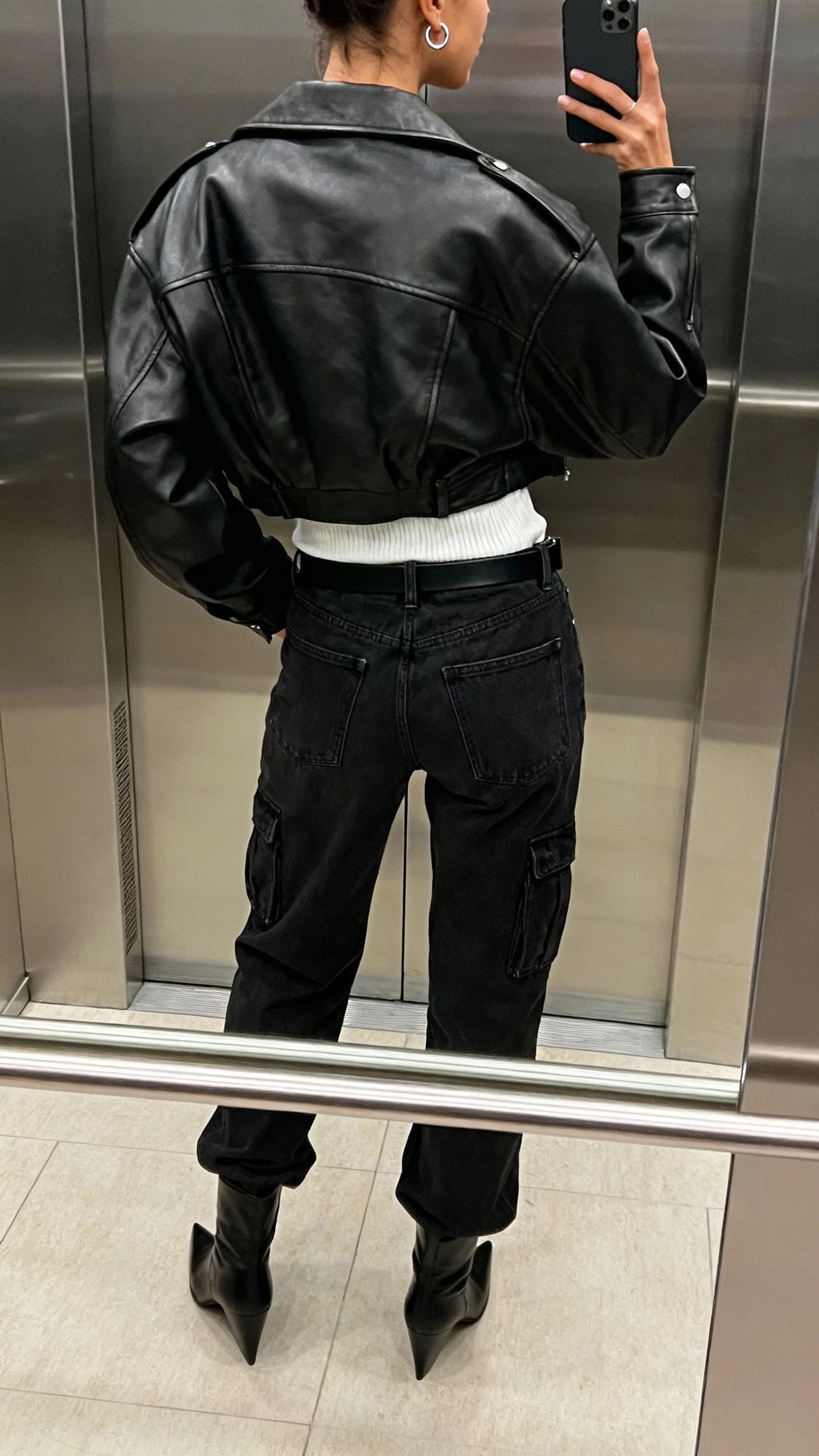 iPhone back-view selfie of a woman wearing black cargo jeans, white ribbed tee, oversized black leather moto jacket, pointed black boots, slim black belt, and small silver hoop earrings; face not visible, elevator mirror, natural indoor lighting, natural iPhone photo quality.