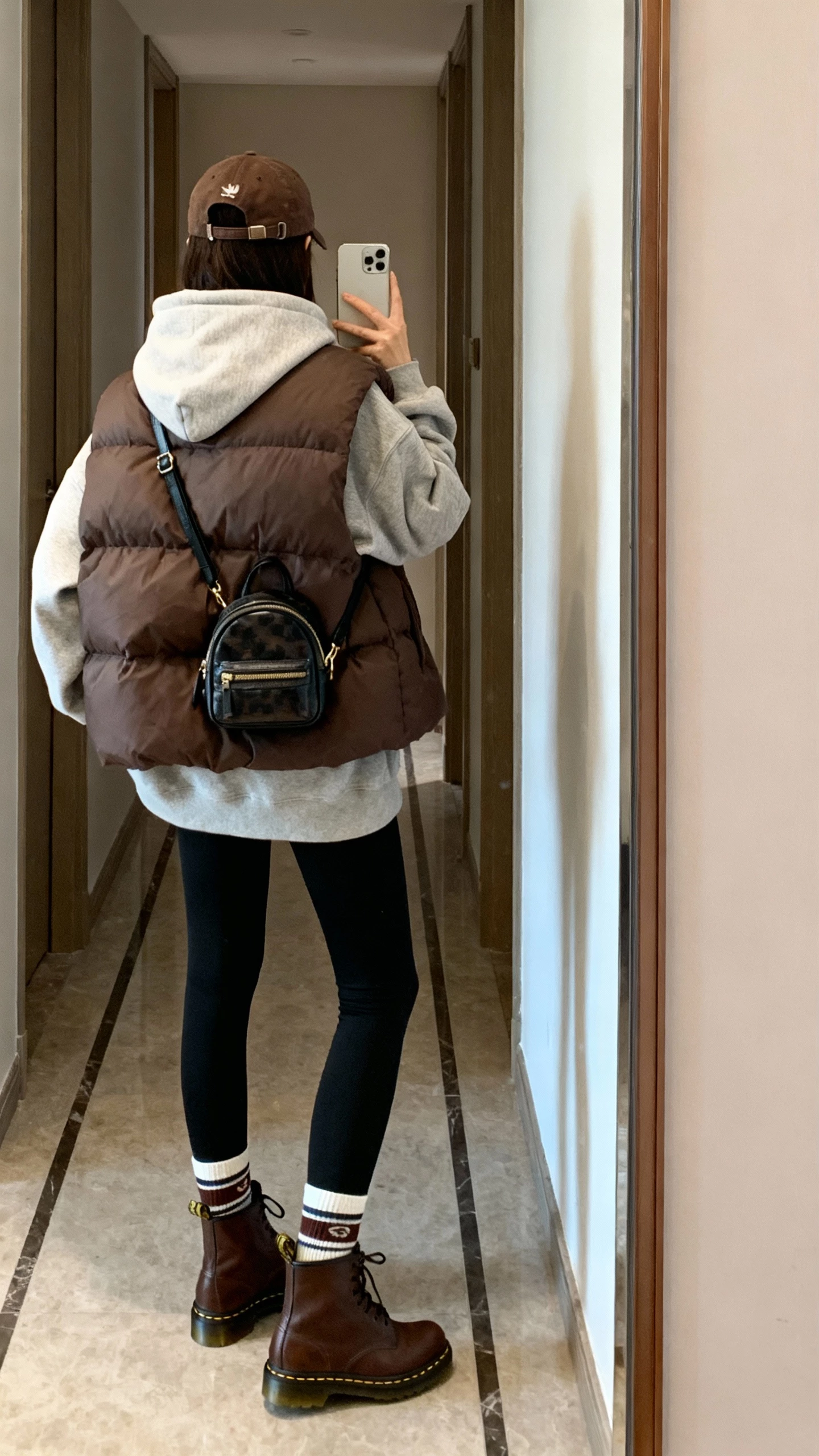 iPhone back-view selfie of a woman wearing brown Doc Martens with black leggings, oversized hoodie, puffer vest, baseball cap, sporty socks peeking, and a mini backpack, face not visible, hallway mirror after a walk, natural daylight, casual iPhone photo.