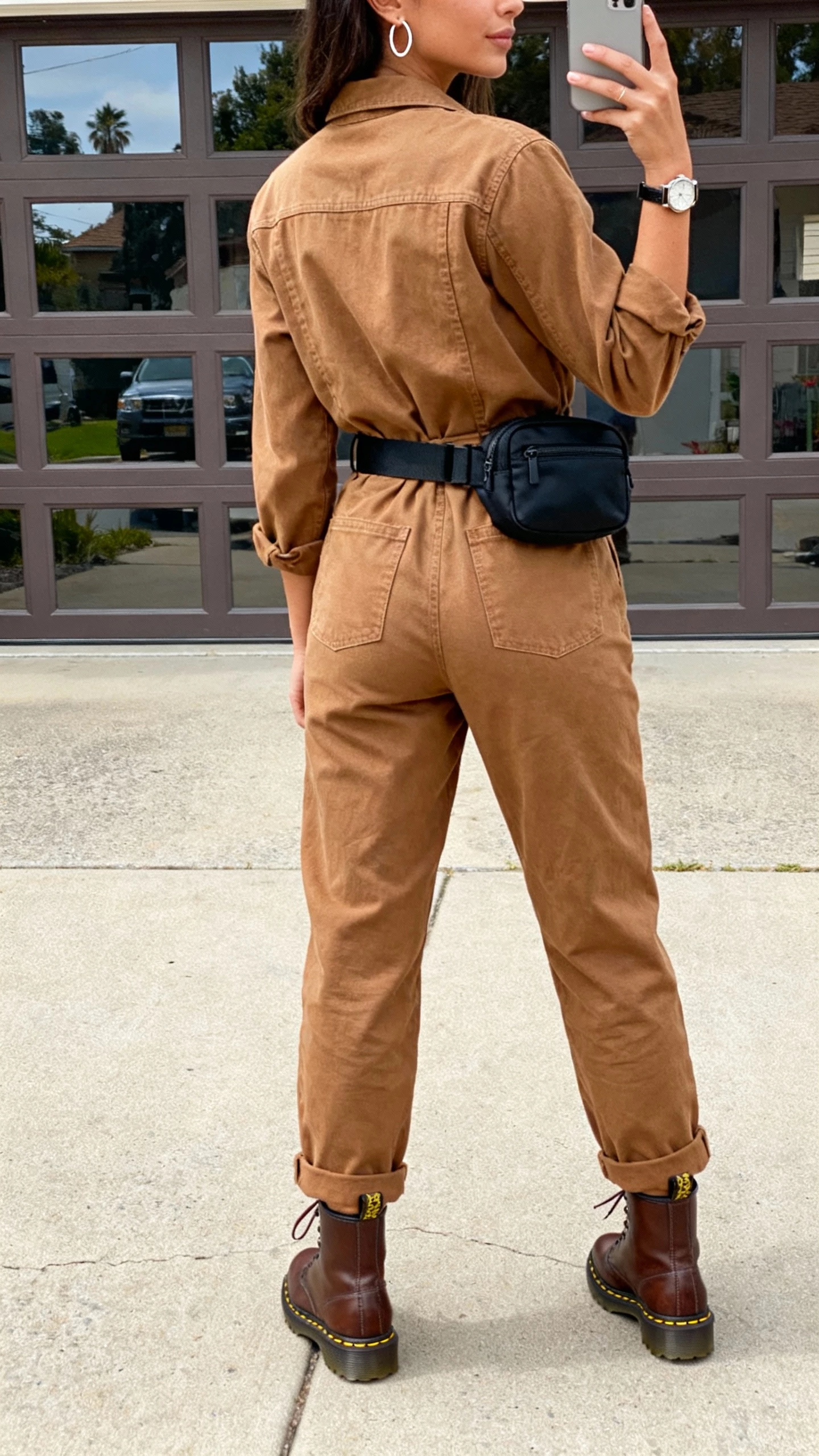 iPhone back-view selfie of a woman wearing brown Doc Martens with a tan utility jumpsuit cinched at the waist, black belt bag, simple hoop earrings, and a watch, face not visible, garage door reflection, afternoon daylight, natural iPhone photo.