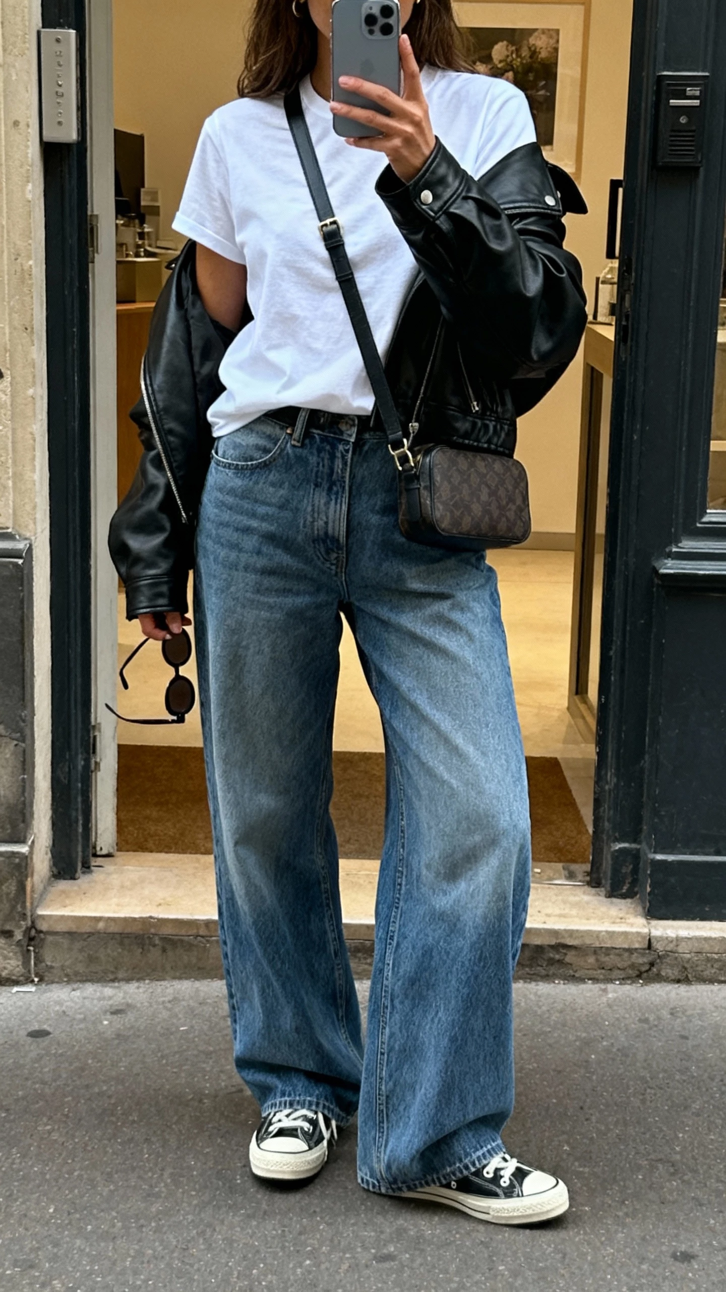 iPhone back-view selfie of a woman wearing wide leg jeans, a relaxed white tee, black leather jacket thrown over shoulders, retro sneakers, crossbody bag and sunglasses in hand, face not visible, city street doorway, natural daylight, casual iPhone vibe.