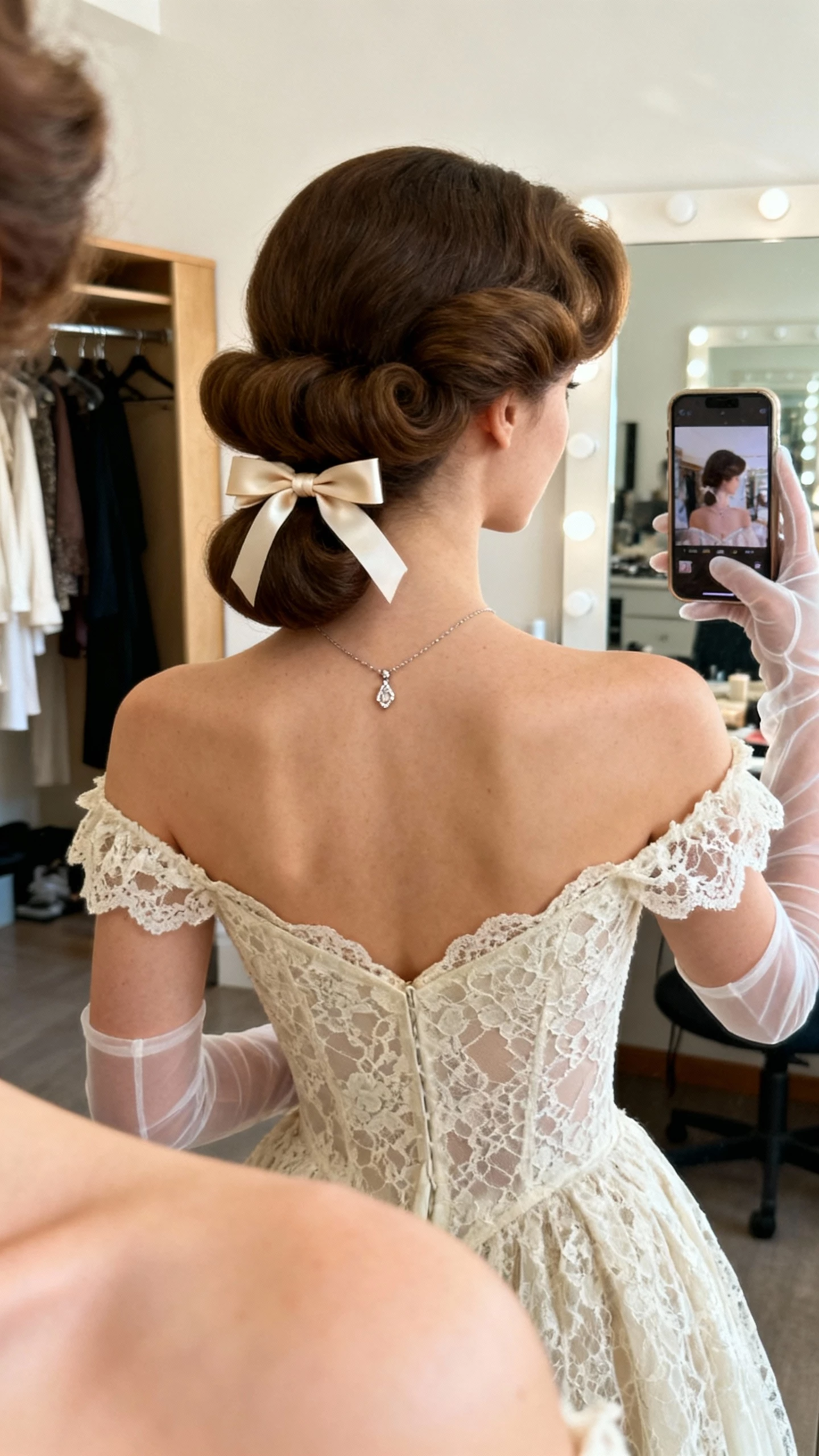 iPhone back-view selfie of a woman with 1960s Bardot half-up volume, sweetheart lace dress, ribbon bow at the half-up, dainty pendant necklace, sheer gloves; face not visible, dressing room mirror, soft morning light, natural iPhone photo quality.