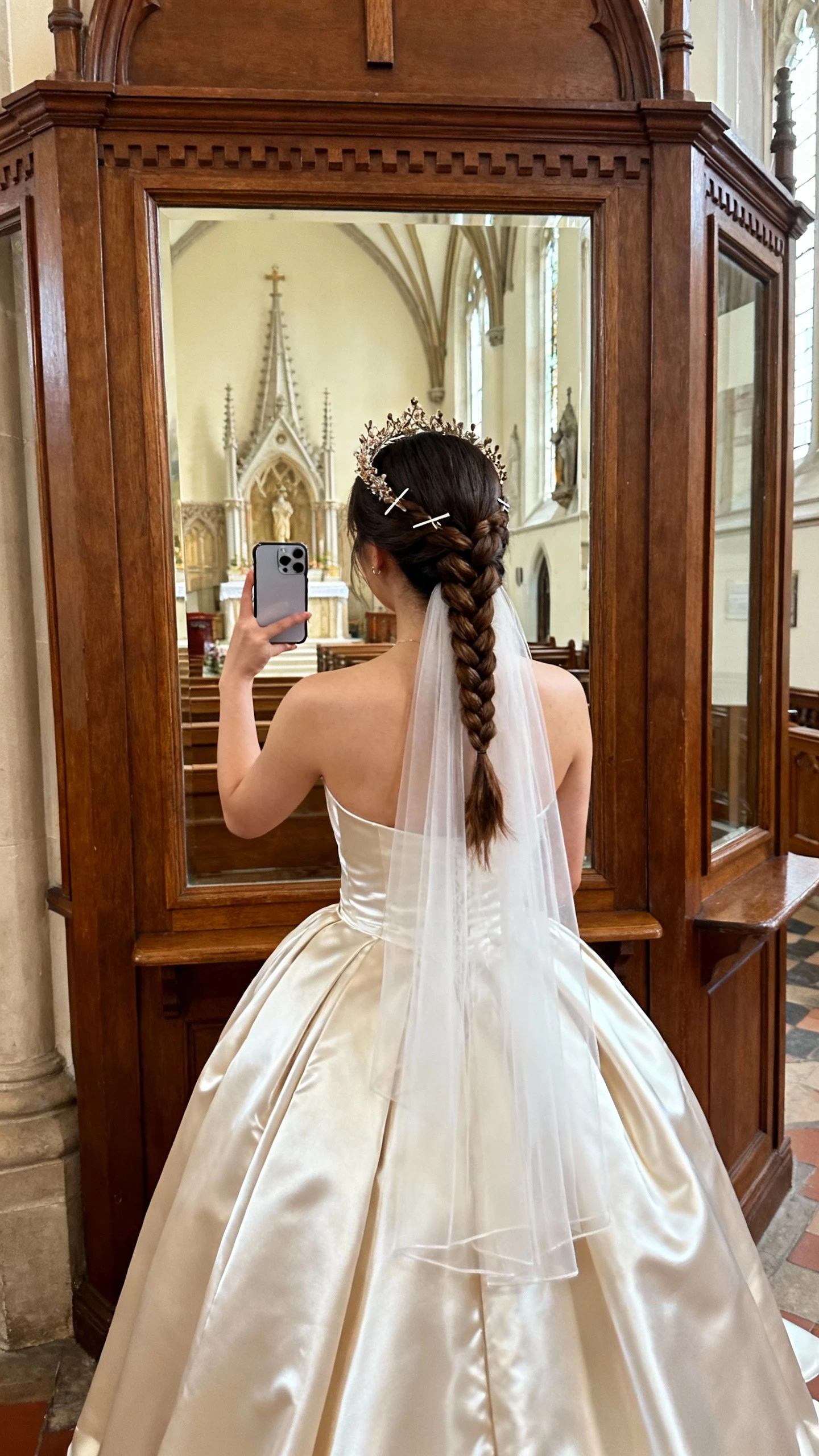 iPhone back-view selfie of a woman with a braided crown, long veil tucked underneath the braid, satin ballgown and delicate hairpins, face not visible, church vestibule mirror, gentle daylight, natural iPhone photo.