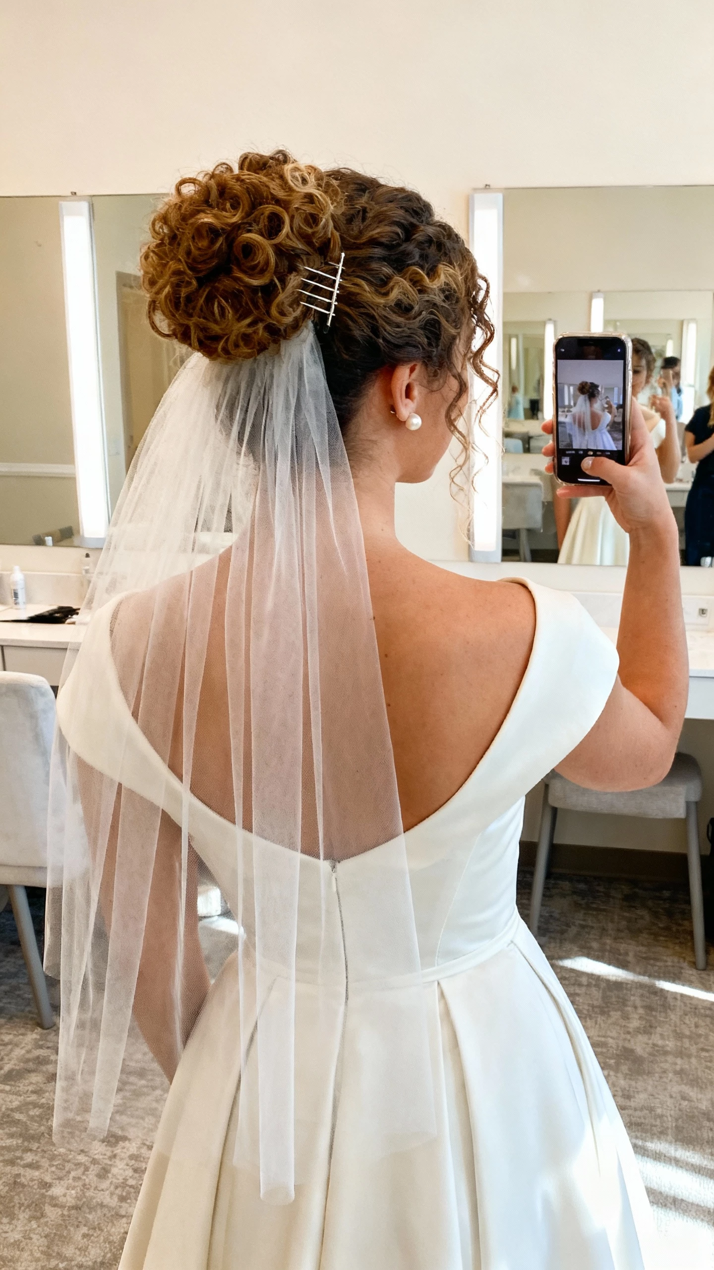 iPhone back-view selfie of a woman with a curly bun featuring veil-friendly comb placement, classic A-line dress, sheer fingertip veil, pearl studs, face not visible, bridal suite mirror, soft morning light, natural iPhone photo.