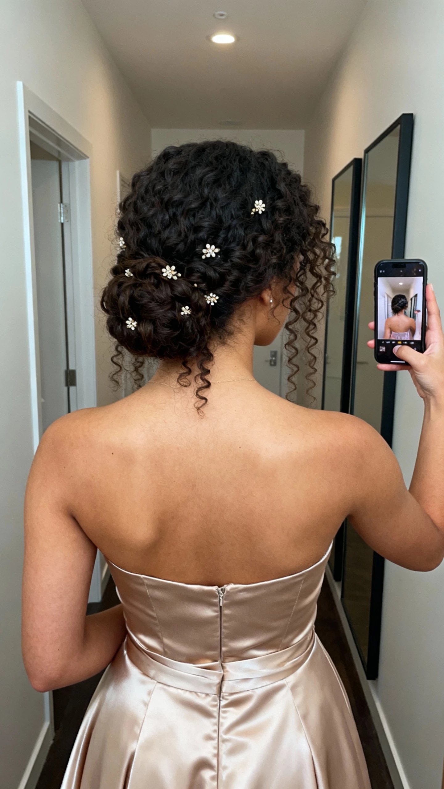 iPhone back-view selfie of a woman with a curly low updo highlighting natural texture, subtle hair jewels, strapless satin gown; face not visible, hallway mirror, soft indoor lighting, natural iPhone photo quality.
