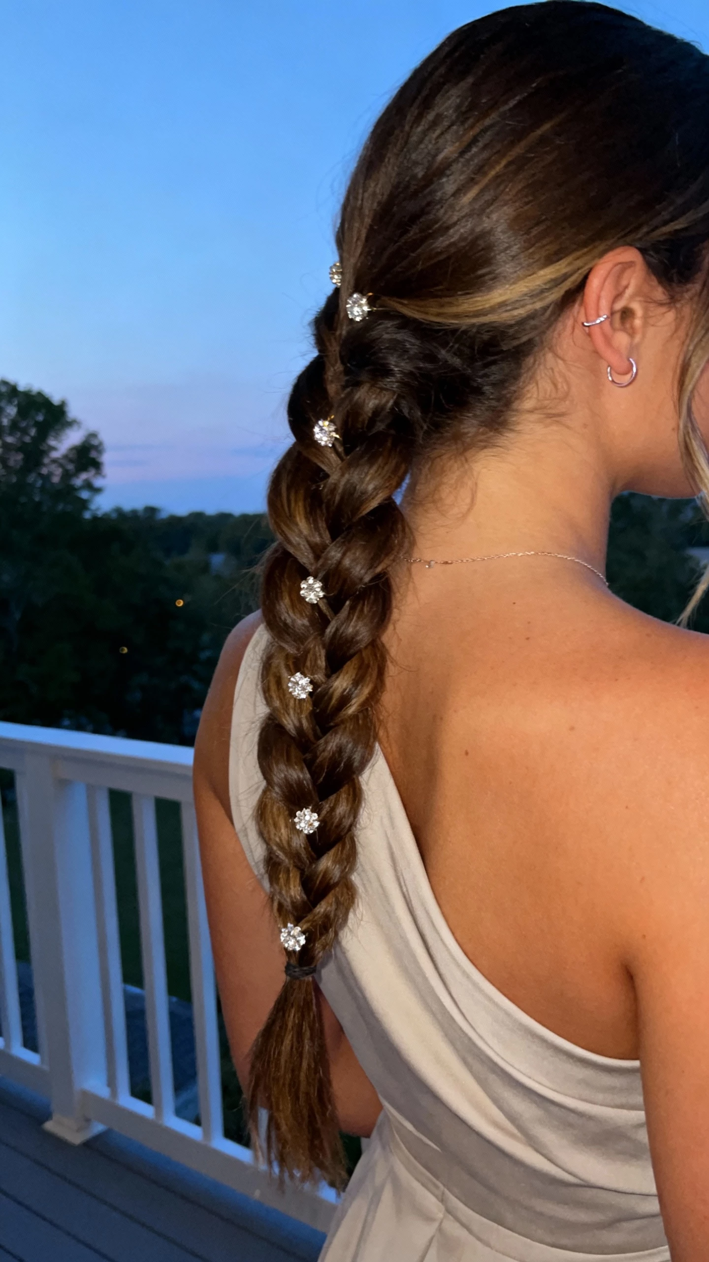 iPhone back-view selfie of a woman with a side-swept fishtail braid adorned with sparkling pins, one-shoulder dress, delicate ear cuff, face not visible, outdoor terrace railing, soft evening light, natural iPhone photo quality.