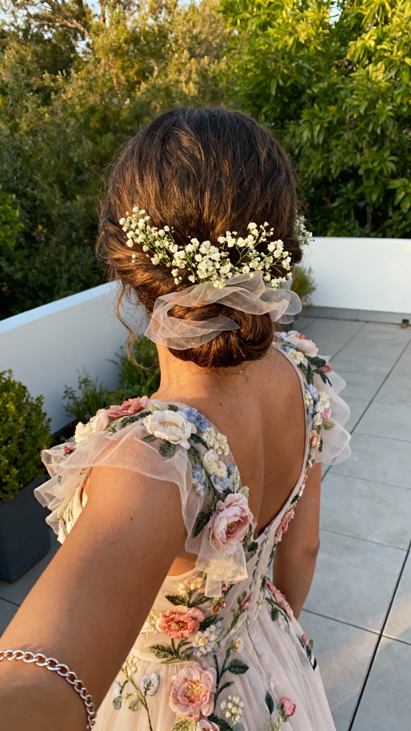 iPhone back-view selfie of a woman with a textured low bun adorned with baby’s breath and whispery layers, romantic garden-style dress with floral appliqués, and delicate chain bracelet; face not visible, outdoor patio near greenery, soft late-afternoon light, natural iPhone aesthetic.