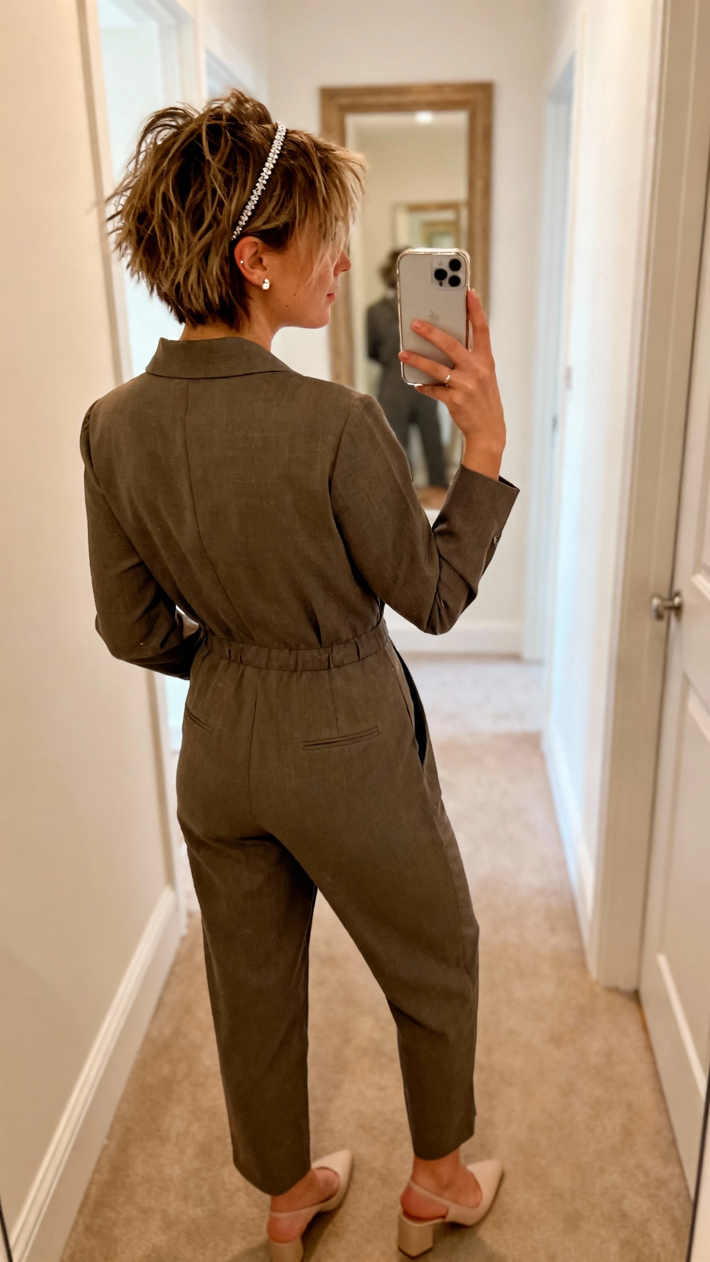 iPhone back-view selfie of a woman with a tousled pixie and a thin sparkly headband, wearing a tailored jumpsuit, dainty stud earrings, and minimalist heels, face not visible, hallway mirror, warm indoor lighting, casual iPhone aesthetic