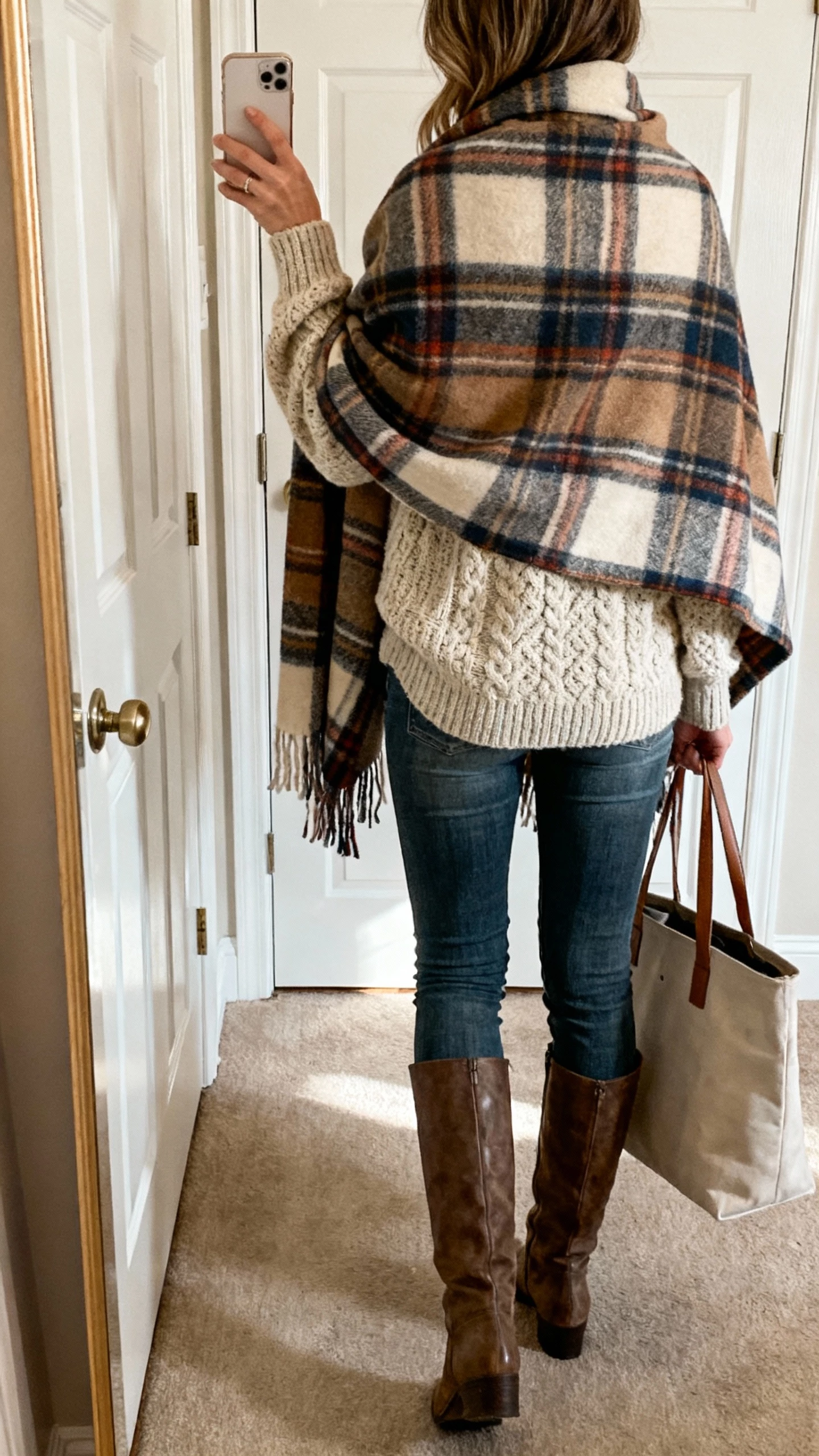 iPhone back-view selfie of a woman wrapped in an oversized plaid blanket scarf worn as a shawl over a chunky knit sweater, skinny jeans, and knee-high boots, tote bag on arm, face not visible, hallway mirror by the door, afternoon natural light, casual iPhone photo.