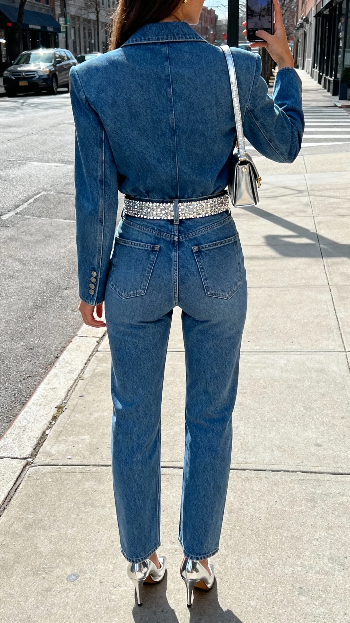 iPhone back-view selfie on a city sidewalk of a woman wearing an elevated denim-on-denim set (tailored denim jacket and high-rise jeans) with a sparkly belt and metallic heels, small shoulder bag, face not visible, natural daylight, casual iPhone vibe