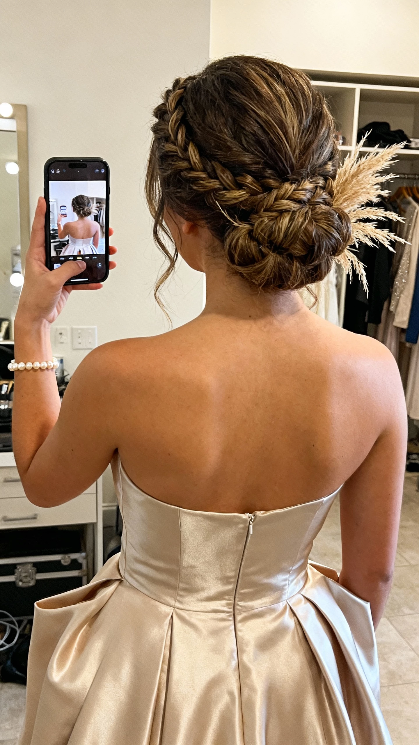 iPhone back-view selfie showing a textured updo with braided details and subtle pampas grass touches, a structured satin gown and pearl bracelet, face not visible, dressing room with warm indoor light, natural iPhone photo quality.