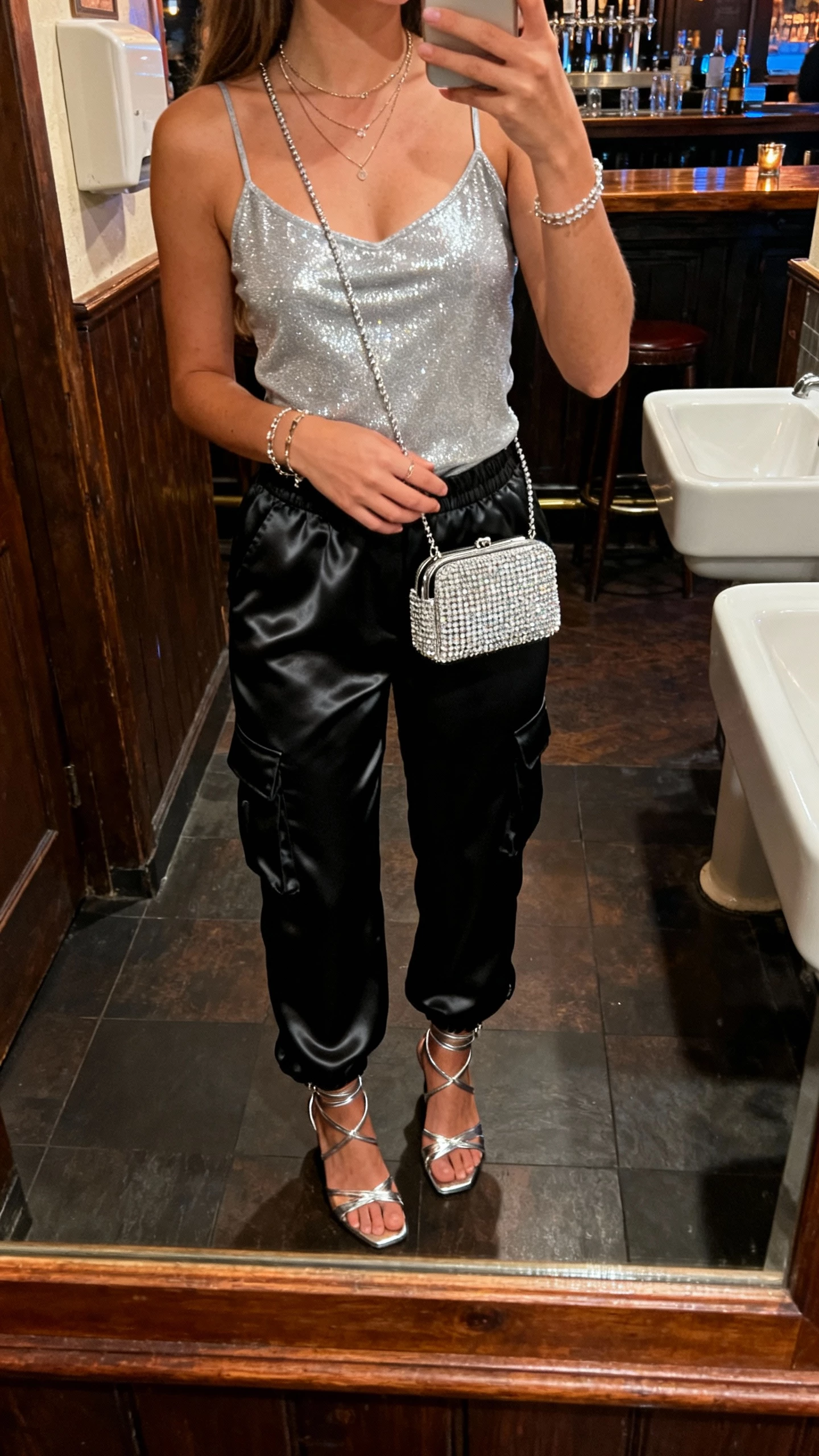 iPhone bar bathroom mirror selfie of a woman wearing satin-finish black cargo pants, sparkly silver camisole, strappy metallic heels, rhinestone mini bag, delicate layered necklaces and bracelets; face not visible, cropped at neck, warm evening indoor lighting, natural iPhone photo quality.