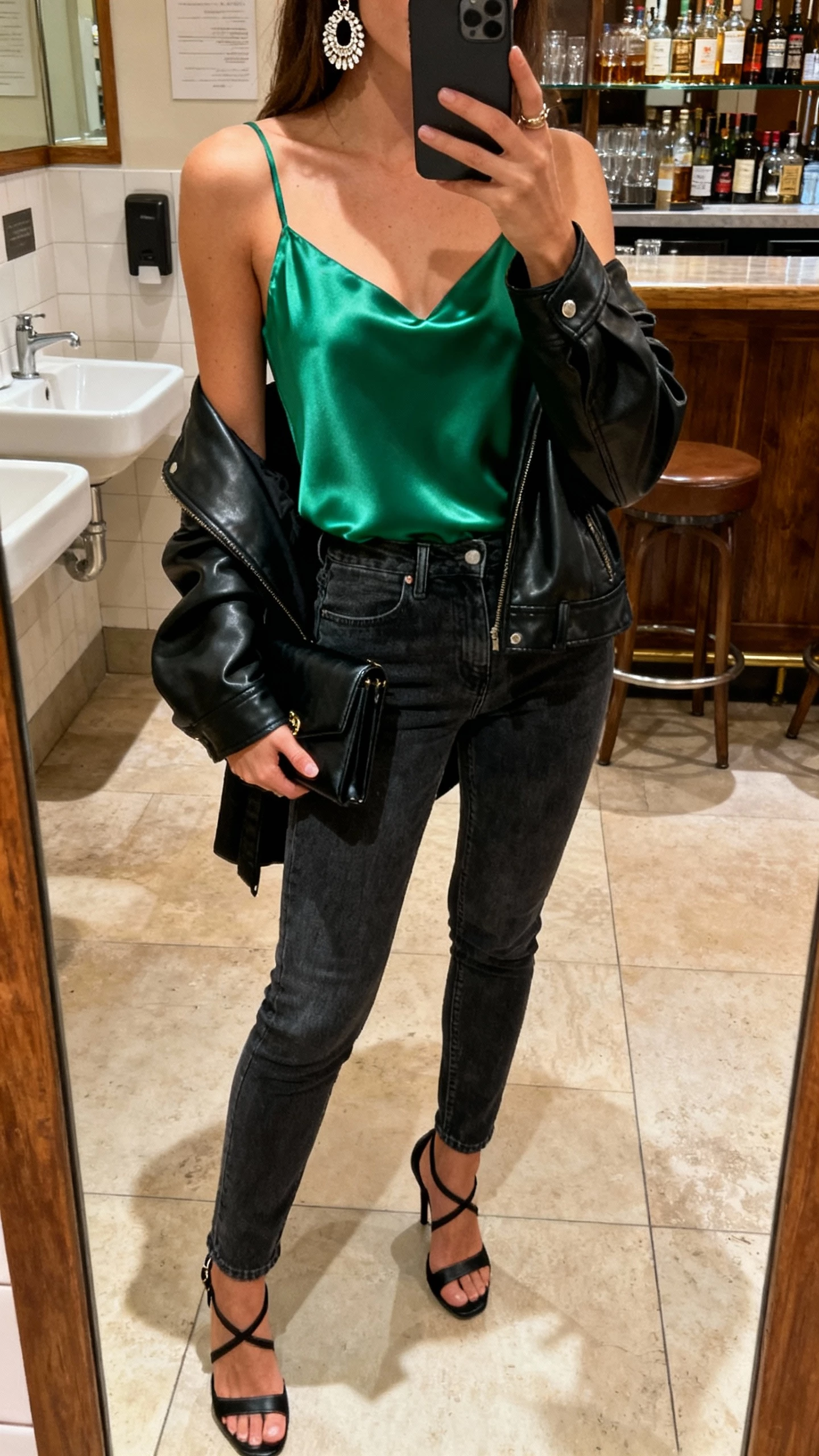 iPhone bar restroom mirror selfie of a woman in an emerald satin camisole, dark-wash slim jeans, black strappy heels, black clutch, statement earrings, leather jacket over shoulder; face not visible, warm indoor lighting, natural iPhone photo quality.