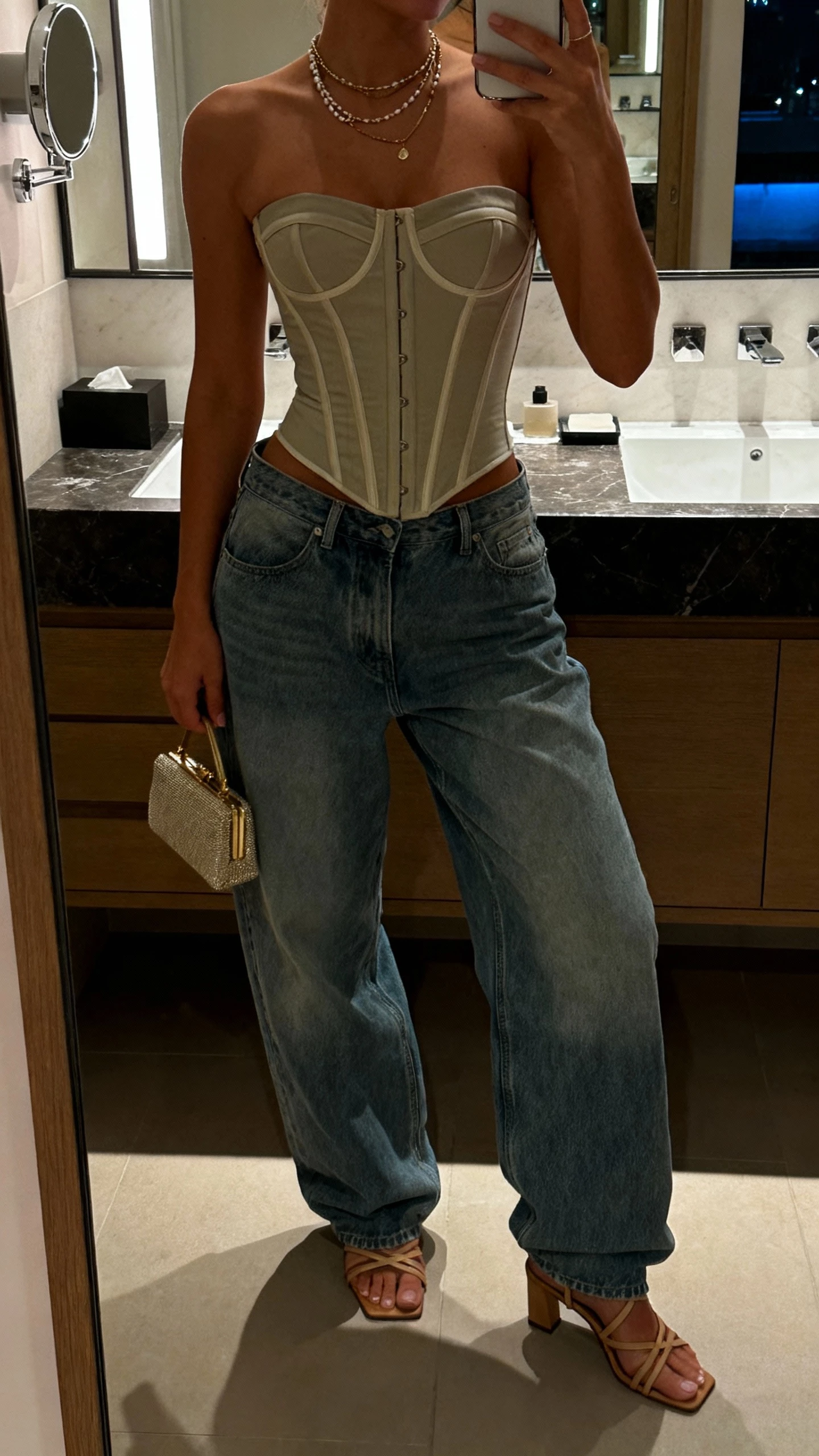 iPhone bathroom mirror selfie of a woman in a structured corset top with baggy denim, strappy heels, petite clutch, layered necklaces; face not visible, cropped at neck, soft evening lighting, natural iPhone photo.