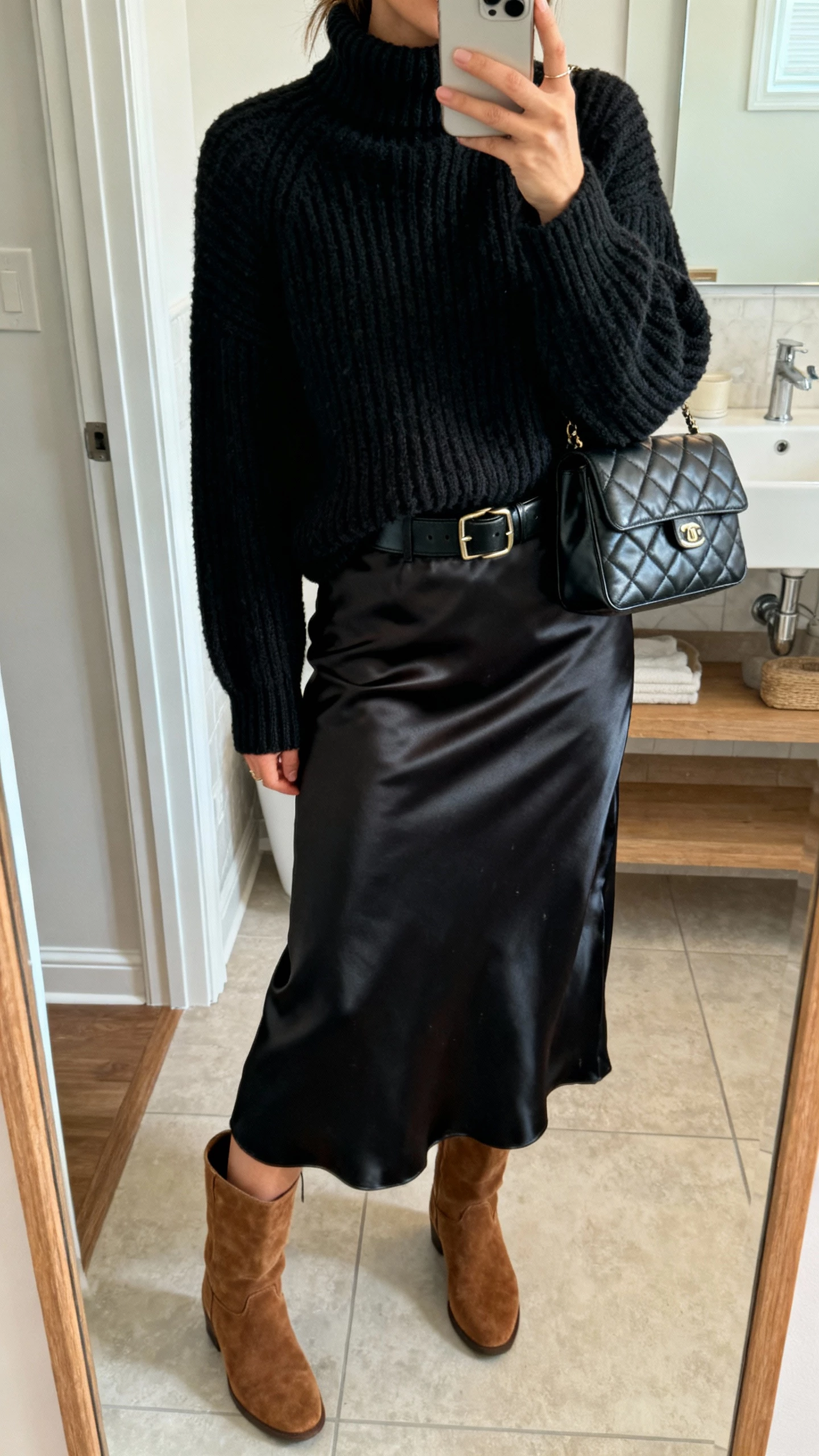iPhone bathroom mirror selfie of a woman in an all-black textured outfit: chunky ribbed turtleneck, satin midi skirt, suede boots, leather belt, and quilted bag, face not visible, cropped at neck, soft indoor lighting, natural iPhone quality.