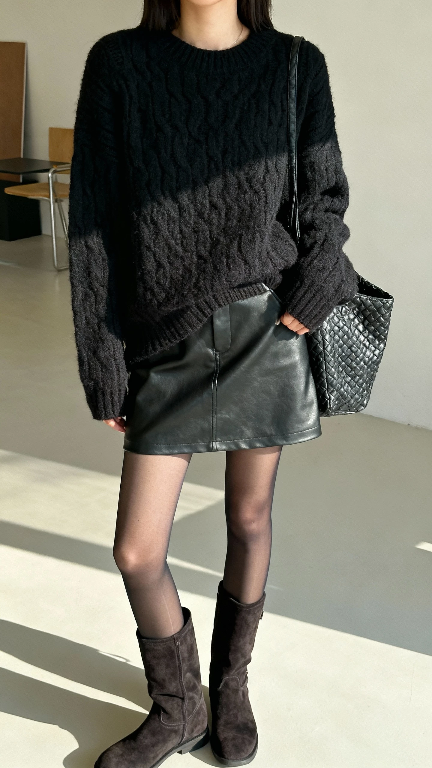 iPhone bathroom mirror selfie of a woman in an all-black look: chunky knit, leather-look skirt, sheer tights, suede boots, and a textured bag, face not visible, cropped at neck, bright indoor light, natural iPhone photo quality.