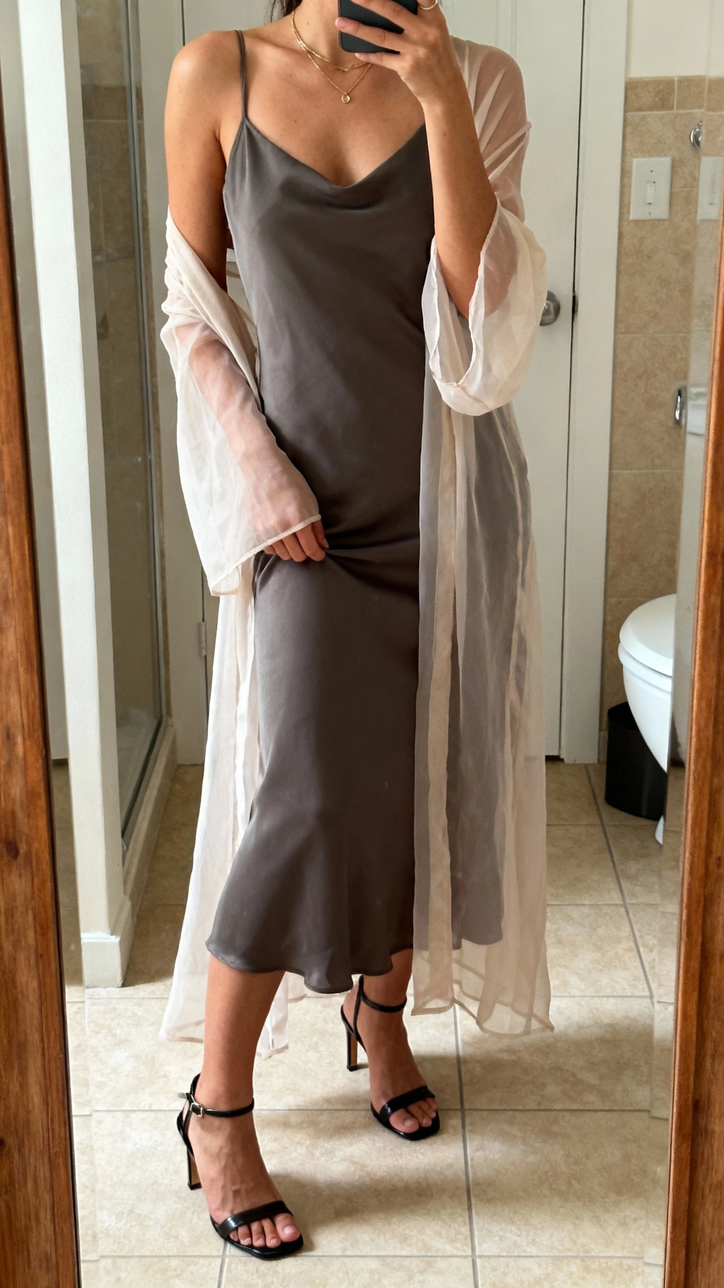 iPhone bathroom mirror selfie of a woman wearing a modern slip dress under a sheer overlay, minimal jewelry, and sleek heeled sandals, face not visible, cropped at shoulders, warm indoor lighting, natural iPhone photo quality.