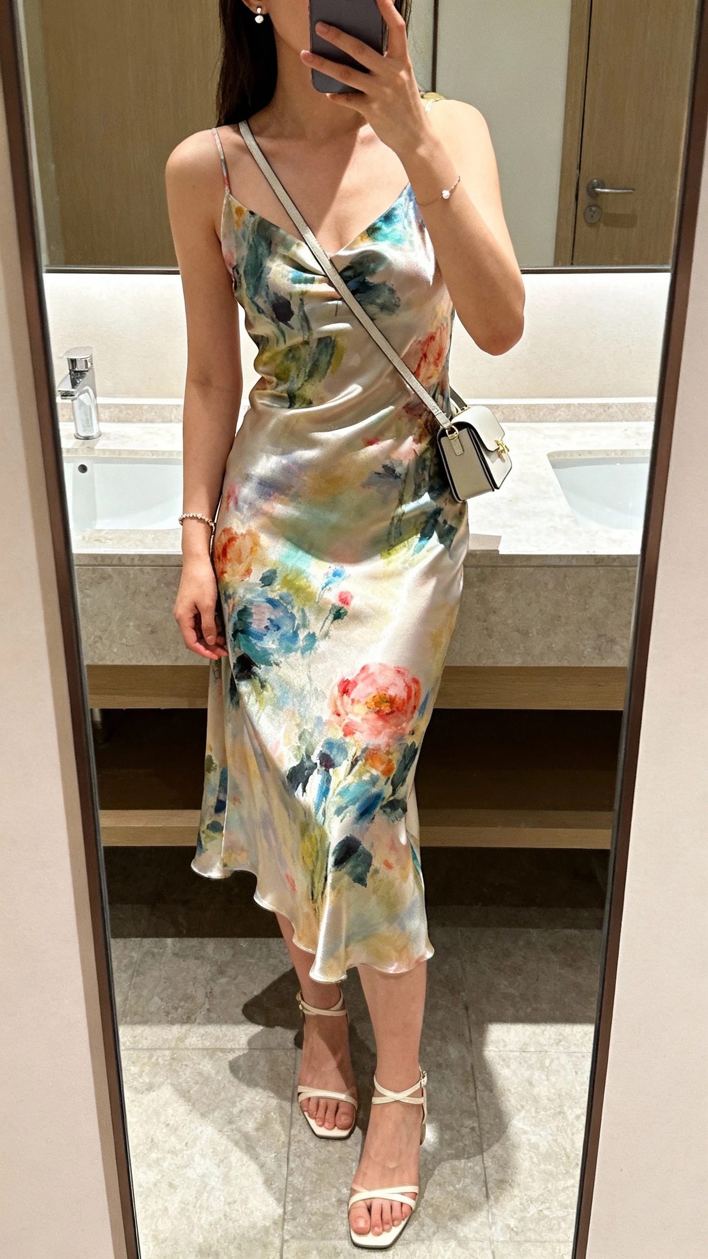 iPhone bathroom mirror selfie of a woman wearing a satin slip dress with painterly floral print, minimal ankle-strap heels, dainty bracelet and tiny stud earrings, sleek mini shoulder bag, face not visible, cropped at shoulders, soft indoor lighting, natural iPhone photo quality