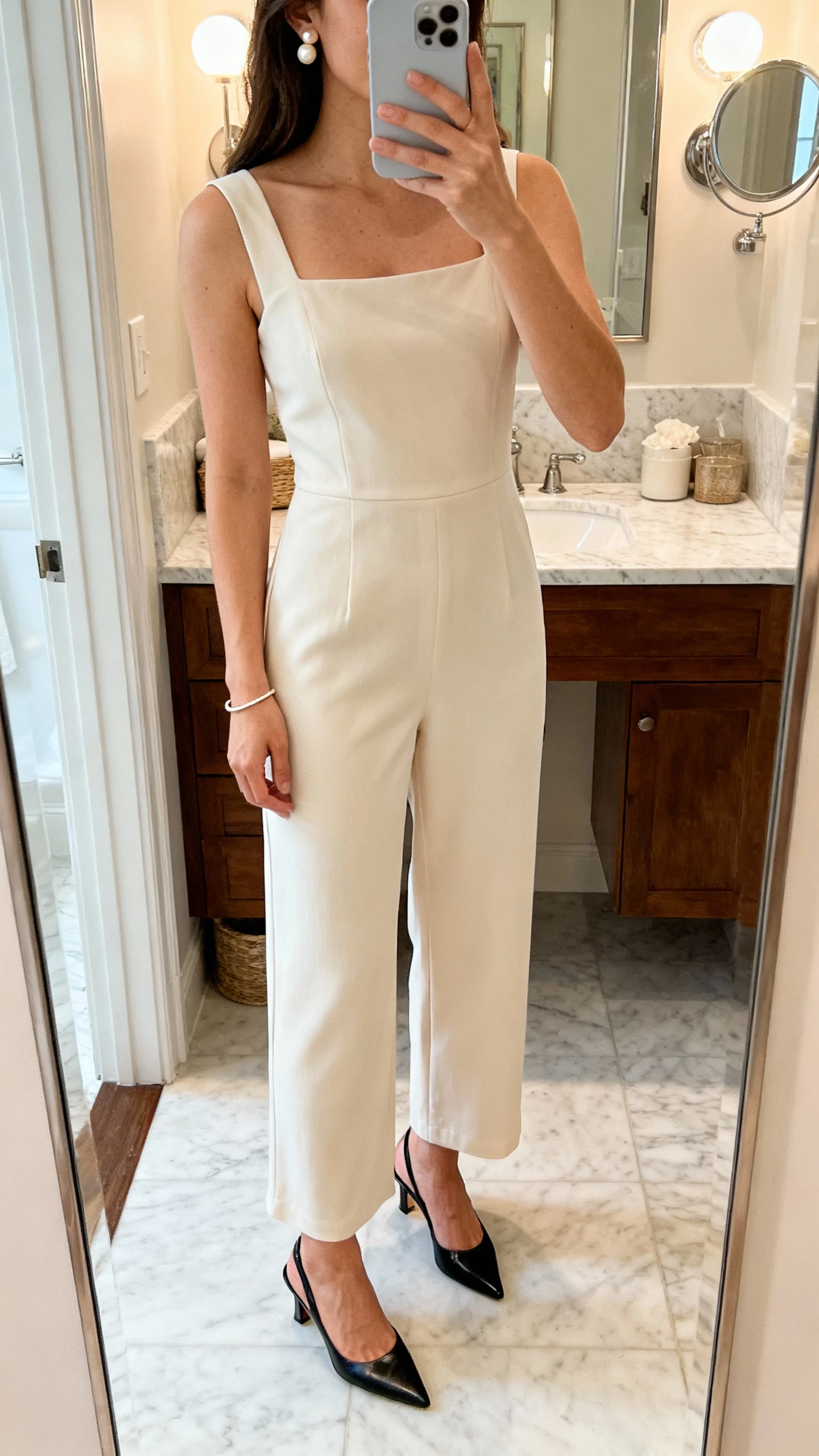 iPhone bathroom mirror selfie of a woman wearing a minimalist square-neck column jumpsuit, sleek slingback heels, pearl studs, thin bracelet, face not visible, soft indoor vanity light, natural iPhone photo quality