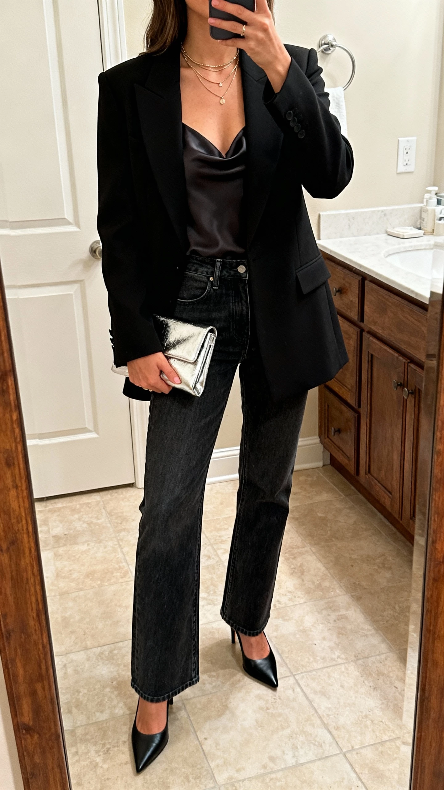 iPhone bathroom mirror selfie of a woman wearing elevated evening denim with a tailored twist: black structured blazer, silky camisole, dark-wash straight jeans, pointed black heels, metallic clutch, delicate layered necklaces; face not visible, cropped at shoulders, warm indoor lighting, natural iPhone photo quality
