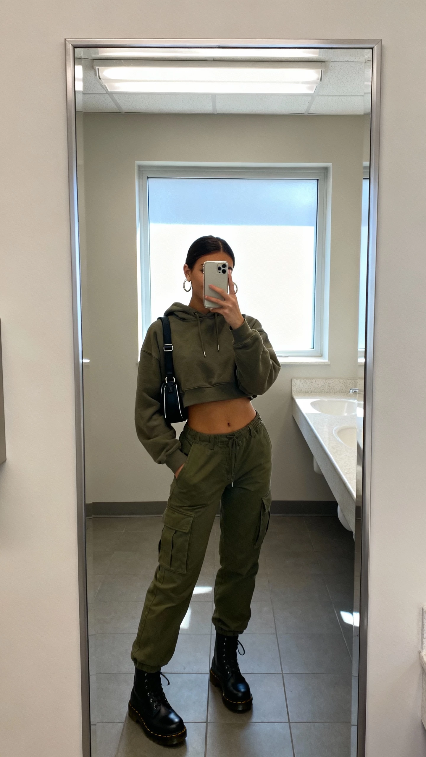 iPhone bathroom mirror selfie of a woman wearing olive cargo pants, a cropped hoodie, black Doc Martens, hoop earrings, a small backpack, face not visible, fluorescent indoor light mixed with window light, casual iPhone photo.