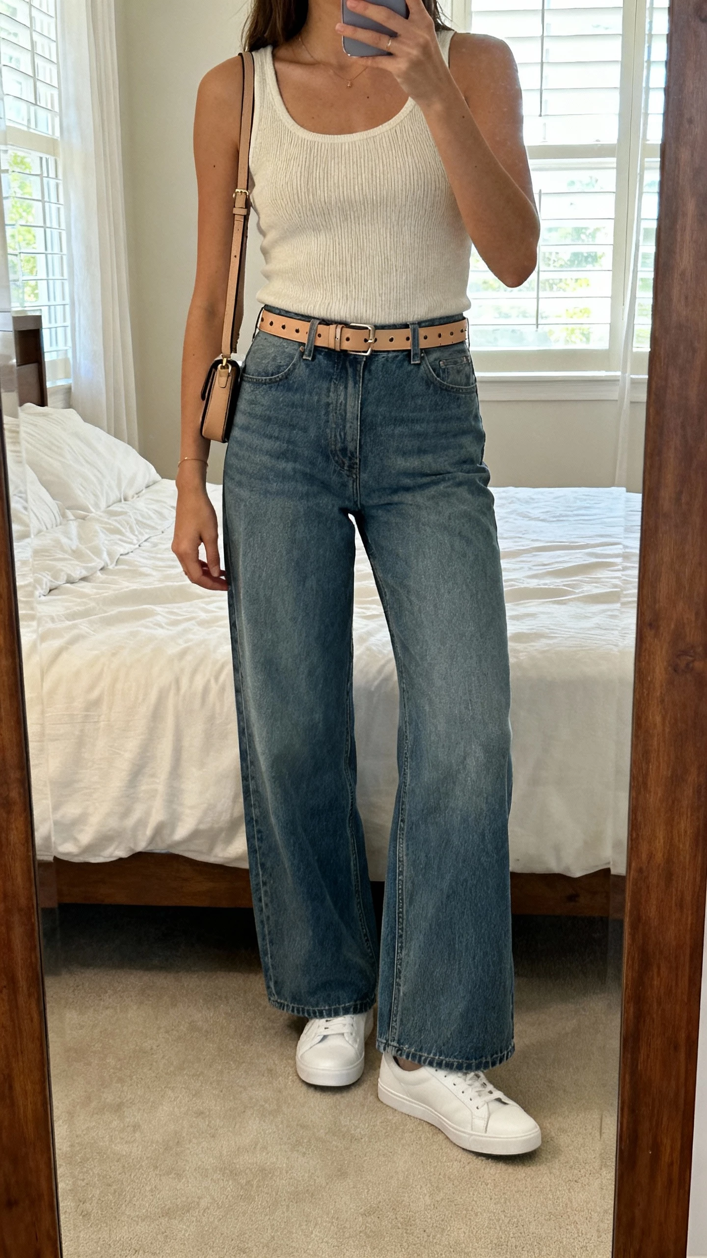 iPhone bedroom mirror selfie of a woman in wide leg jeans, minimalist knit tank, clean white sneakers, slim leather belt, subtle studs, small crossbody, face not visible, soft natural window light, natural iPhone photo quality.