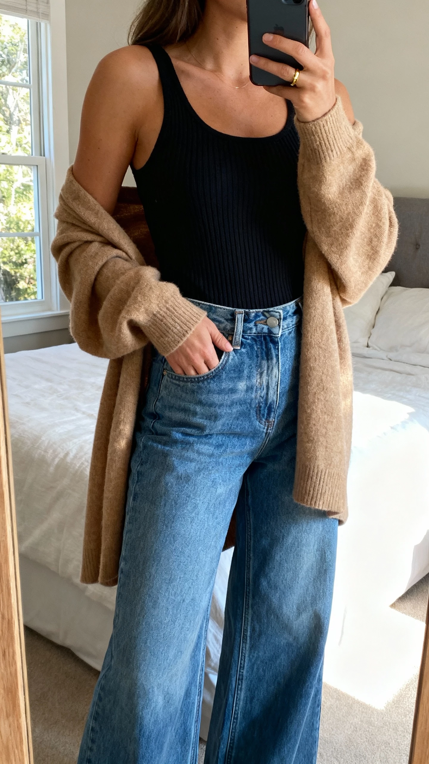 iPhone bedroom mirror selfie of a woman wearing a ribbed black tank, soft camel cardigan draped open, high-rise wide-leg blue jeans, minimal gold ring, face not visible, gentle window light, natural iPhone aesthetic.
